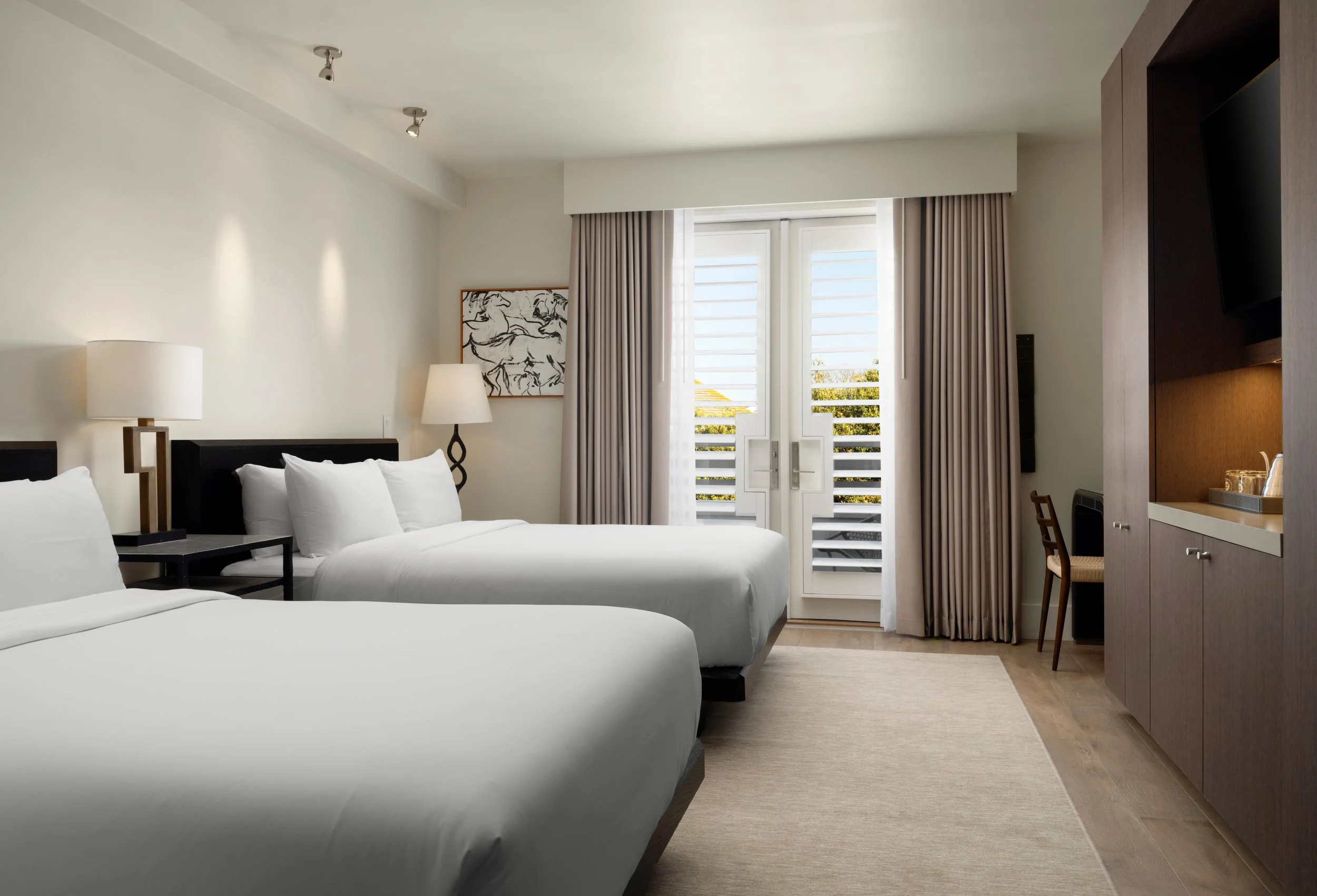 A hotel room with two beds, bedside tables, lamps, a window with curtains, a framed artwork of horses, and a built-in desk area.