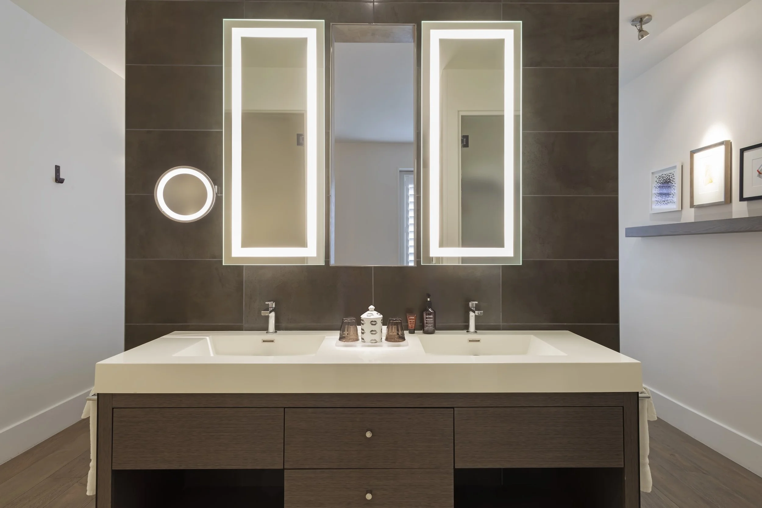 A modern bathroom vanity with a large mirror with LED backlighting, a white countertop with two sinks, and dark wood cabinetry. Framed artwork hangs on the wall on the right, and a small, illuminated mirror is on the left wall.