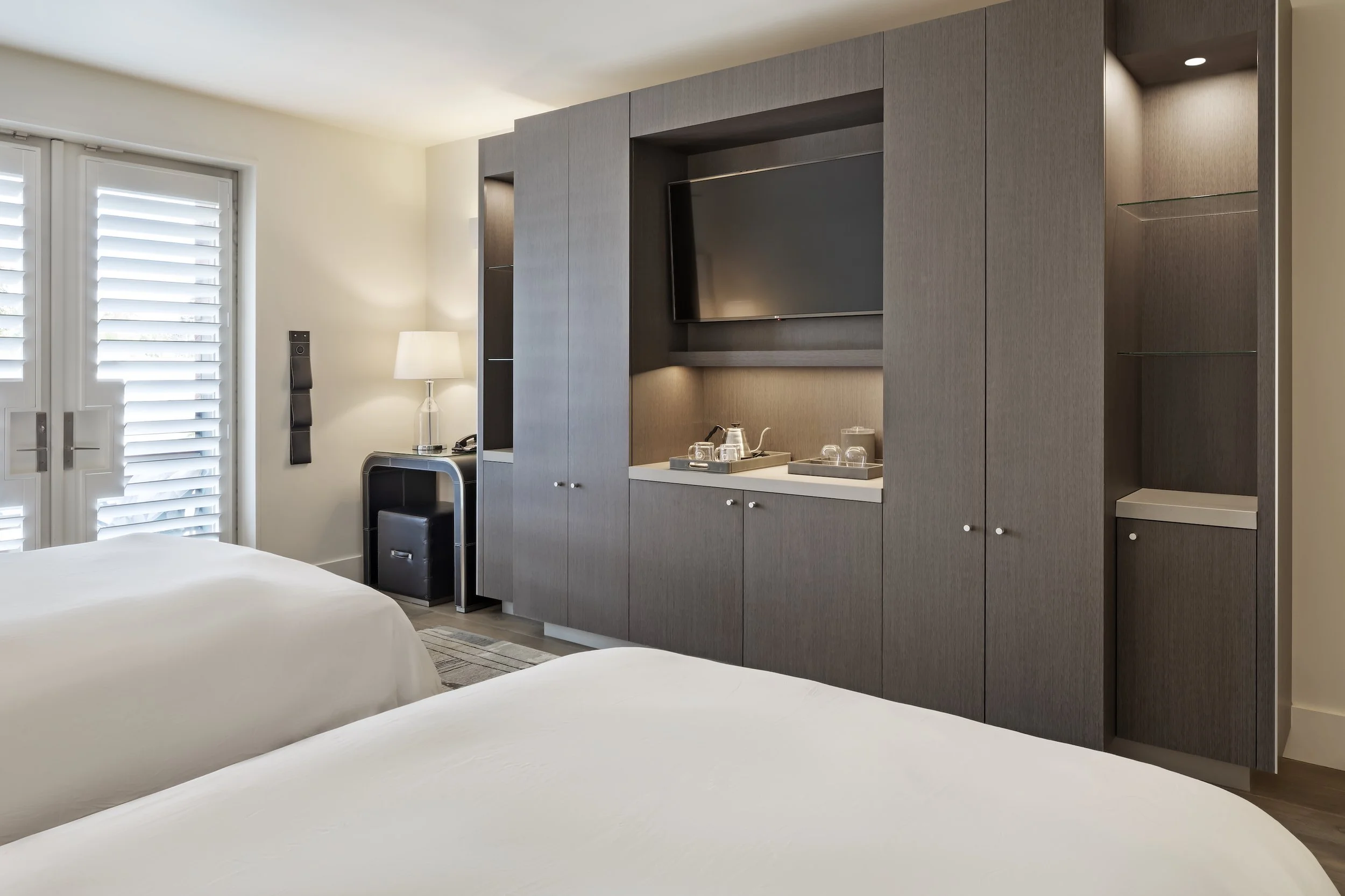 Modern hotel room with two white beds, a wall-mounted TV, a built-in cabinet, and a door with white shutters.