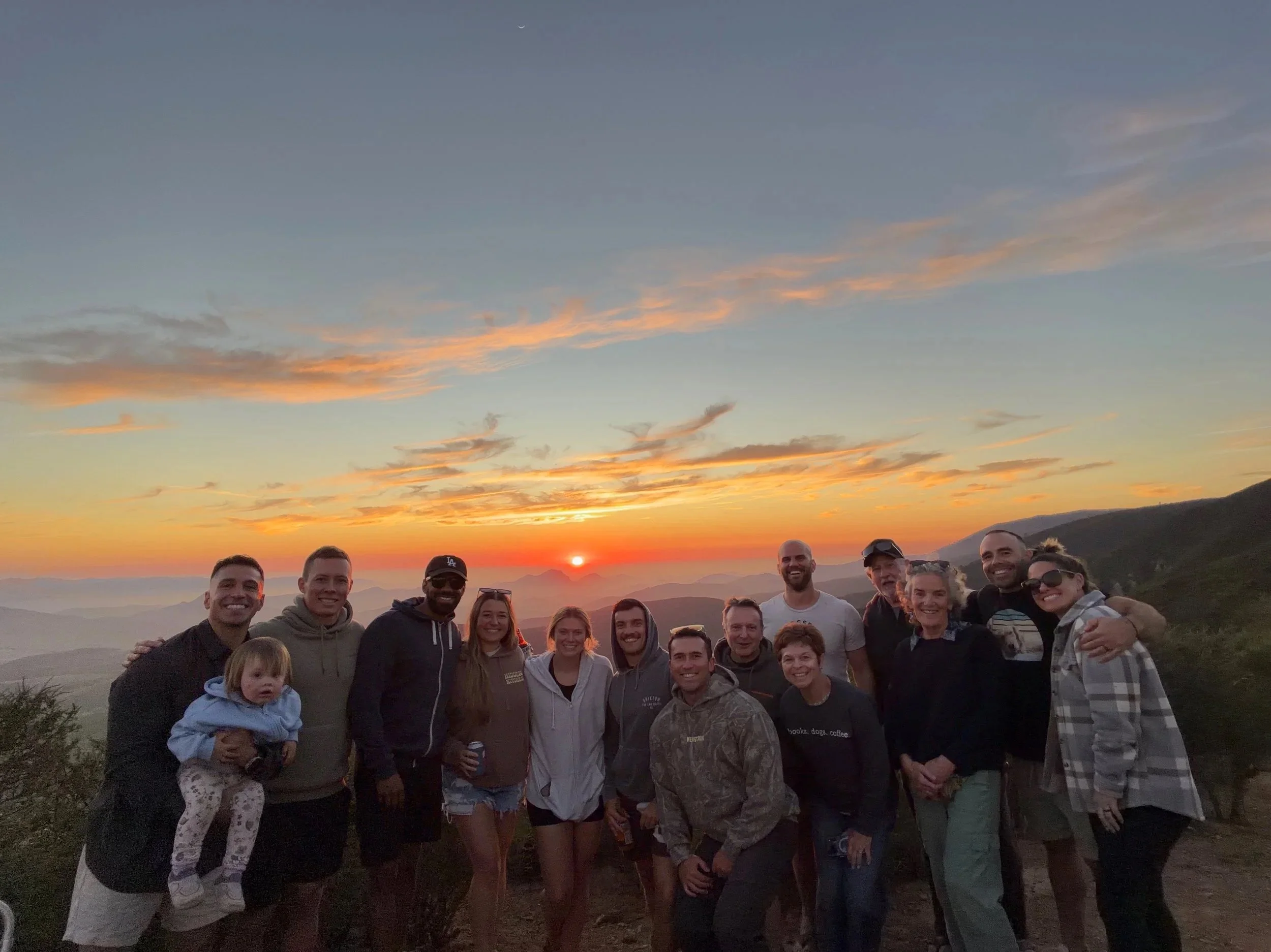 A group of smiling people stands outdoors at sunset, with mountains in the background and a green landscape.