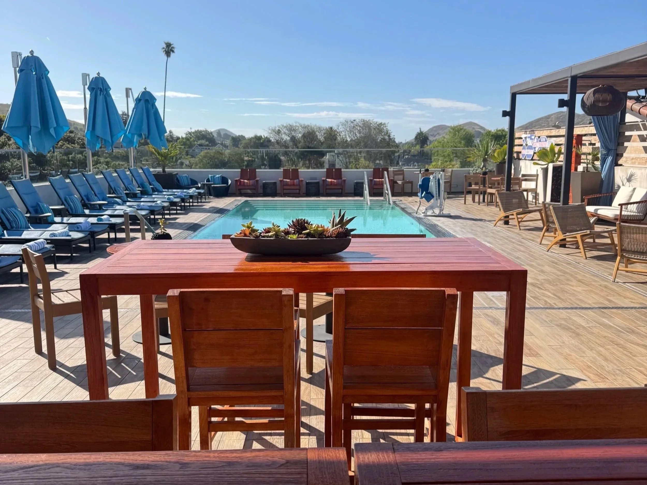 Rooftop pool area with blue umbrellas, sun loungers, and outdoor seating, surrounded by a glass railing and scenic mountain views in the background.