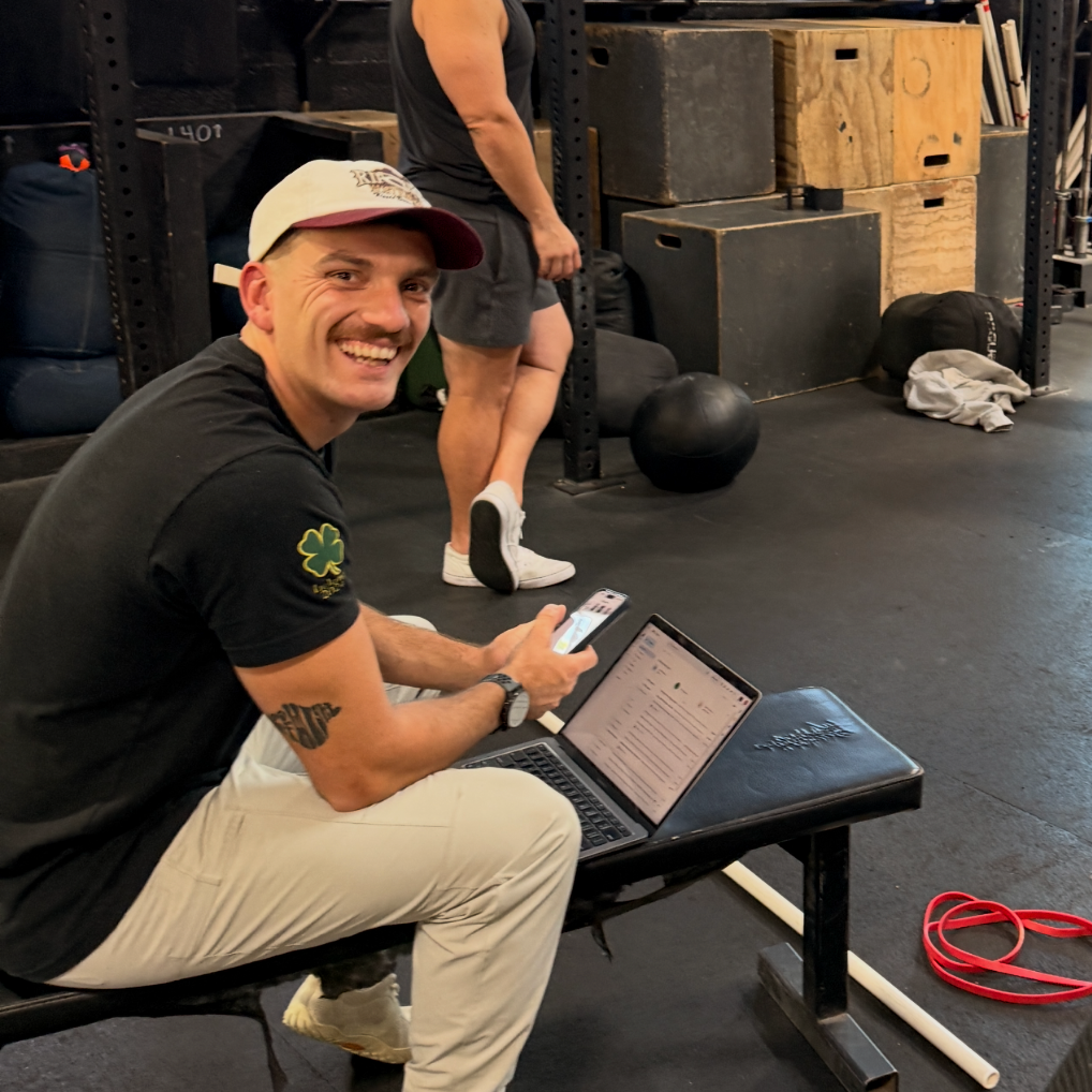 A man smiling while sitting on a bench in a gym, with a laptop and phone, and another person in the background near gym equipment.