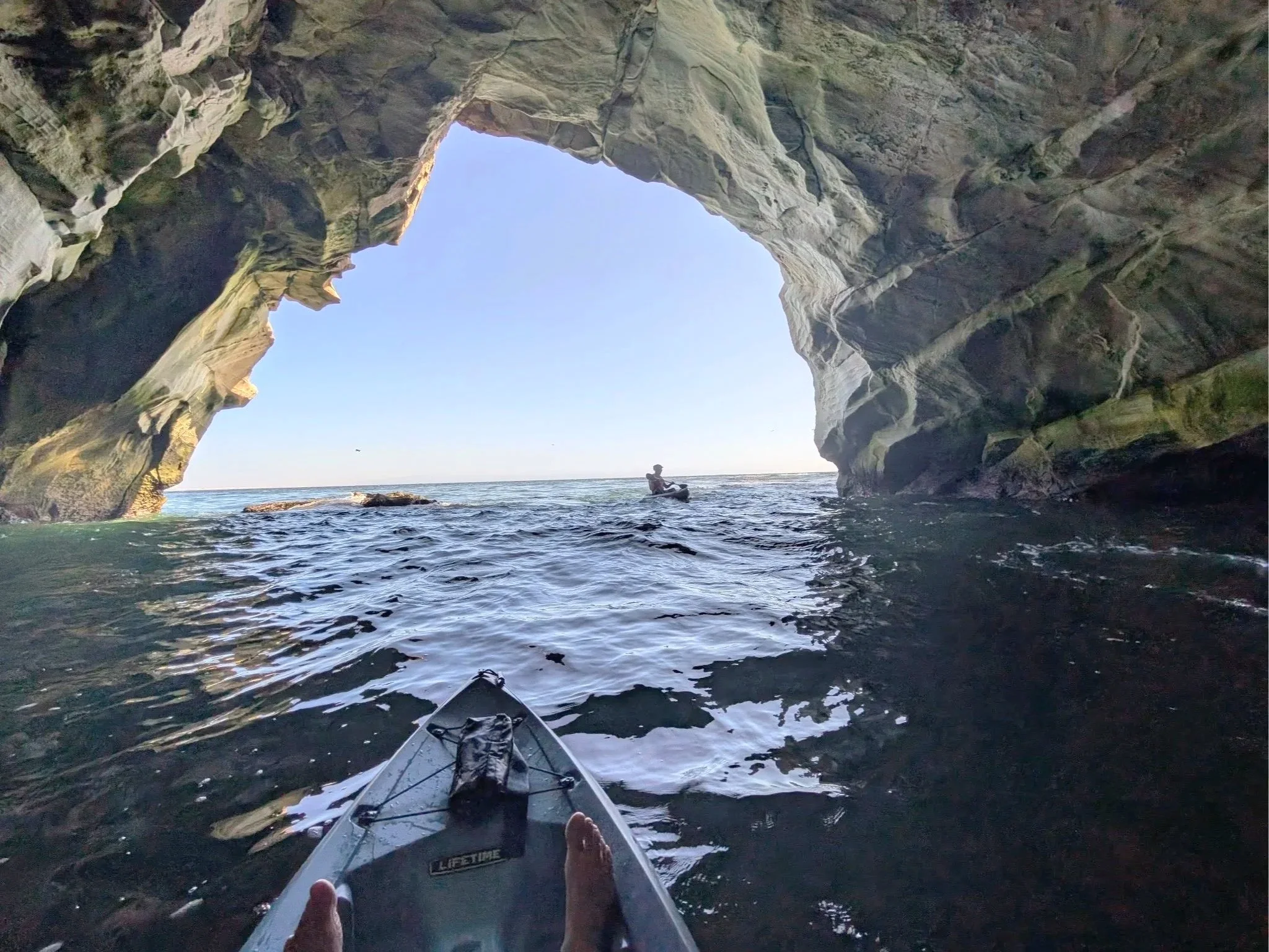 View from a kayak inside a large sea cave, with a person kayaking near the rocky cave walls and an opening to the sky seen at the top.