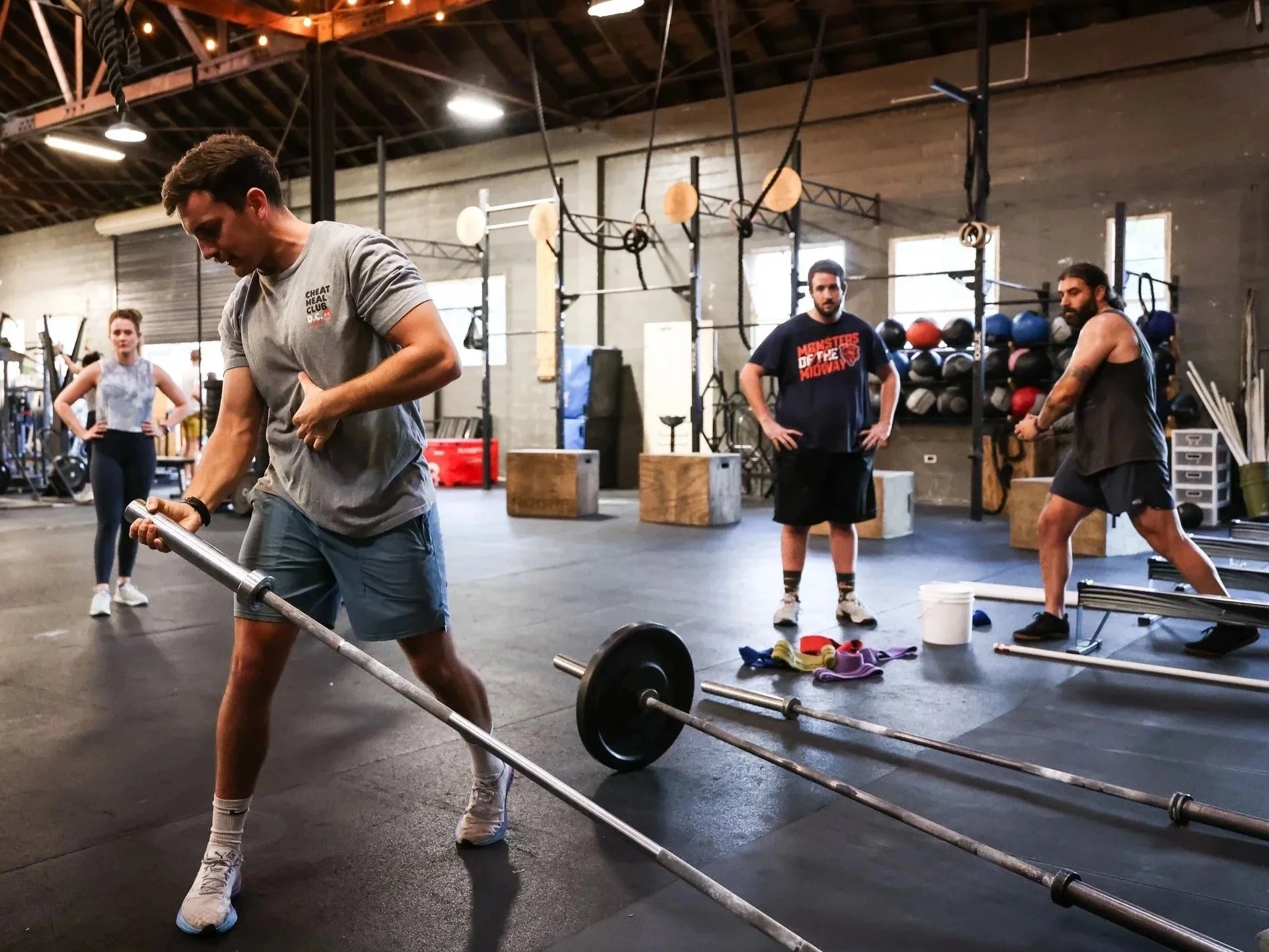 A person lifting a barbell in a gym with other people in the background. The gym is spacious with high ceilings, wooden beams, and various workout equipment.