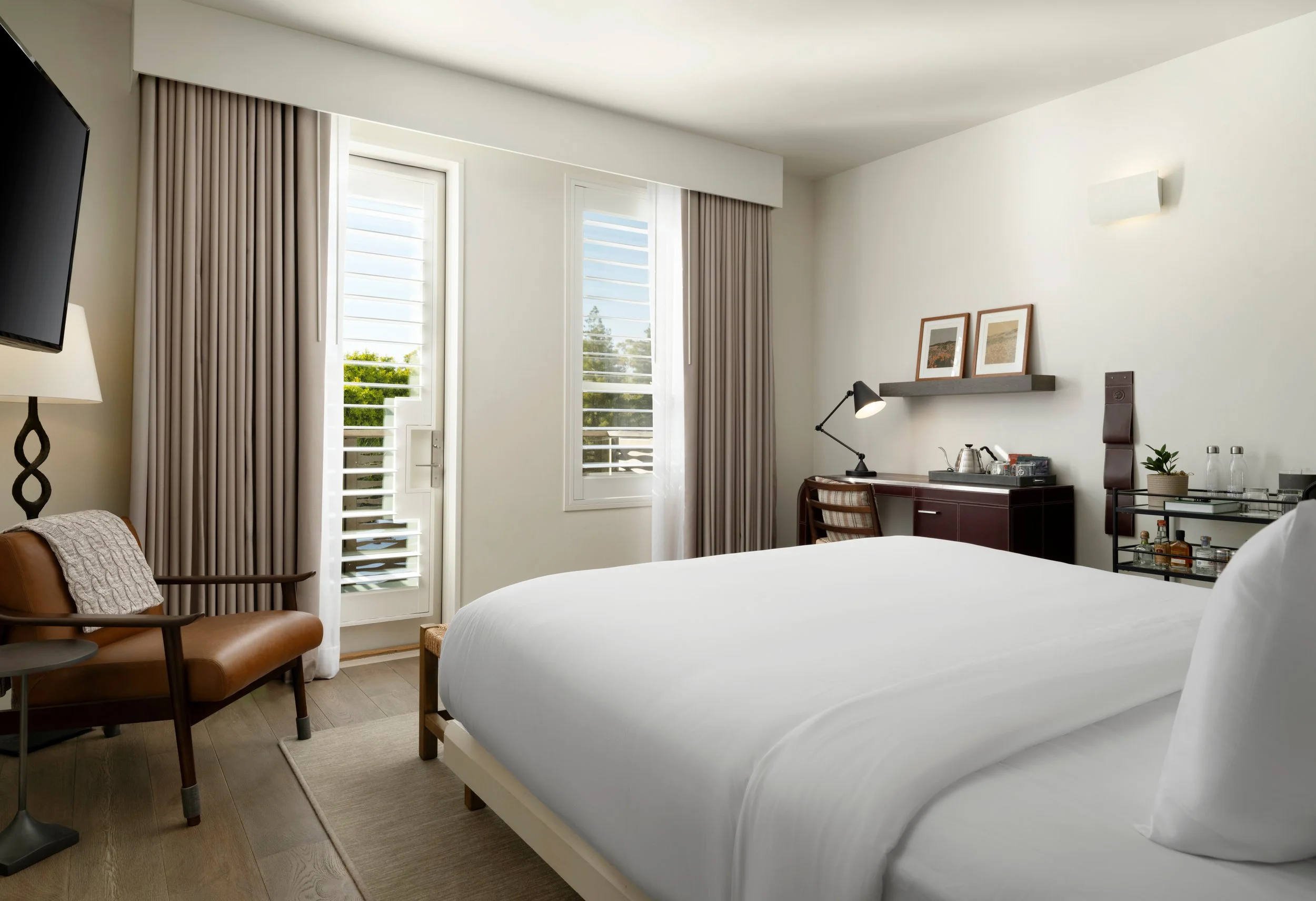 A modern hotel room with large windows, beige curtains, a white bed, a leather armchair, a small desk with a lamp, framed artwork, and a minibar.