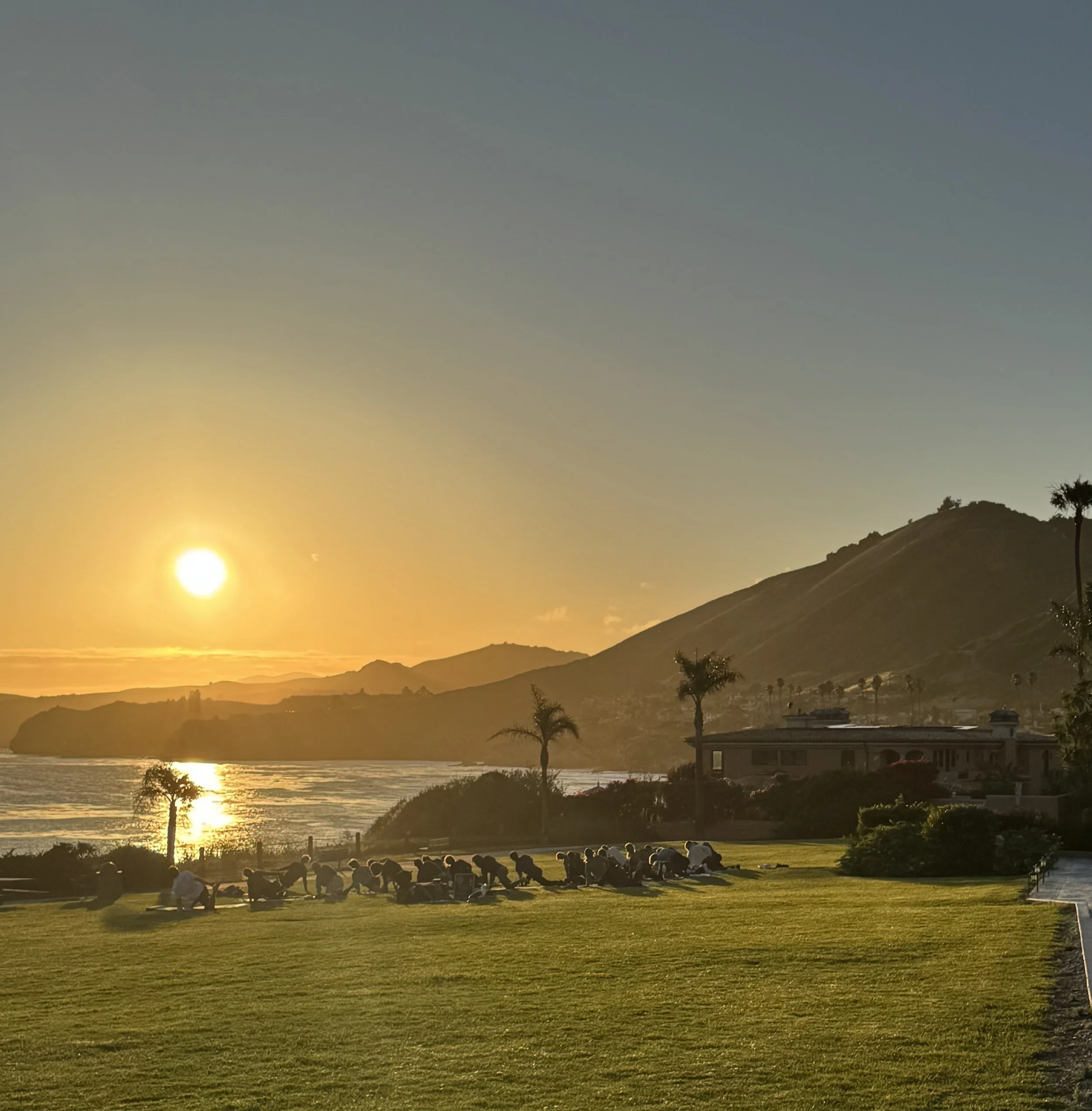 Sunset over a body of water with mountains and a clear sky, people sitting on a grassy area in foreground.