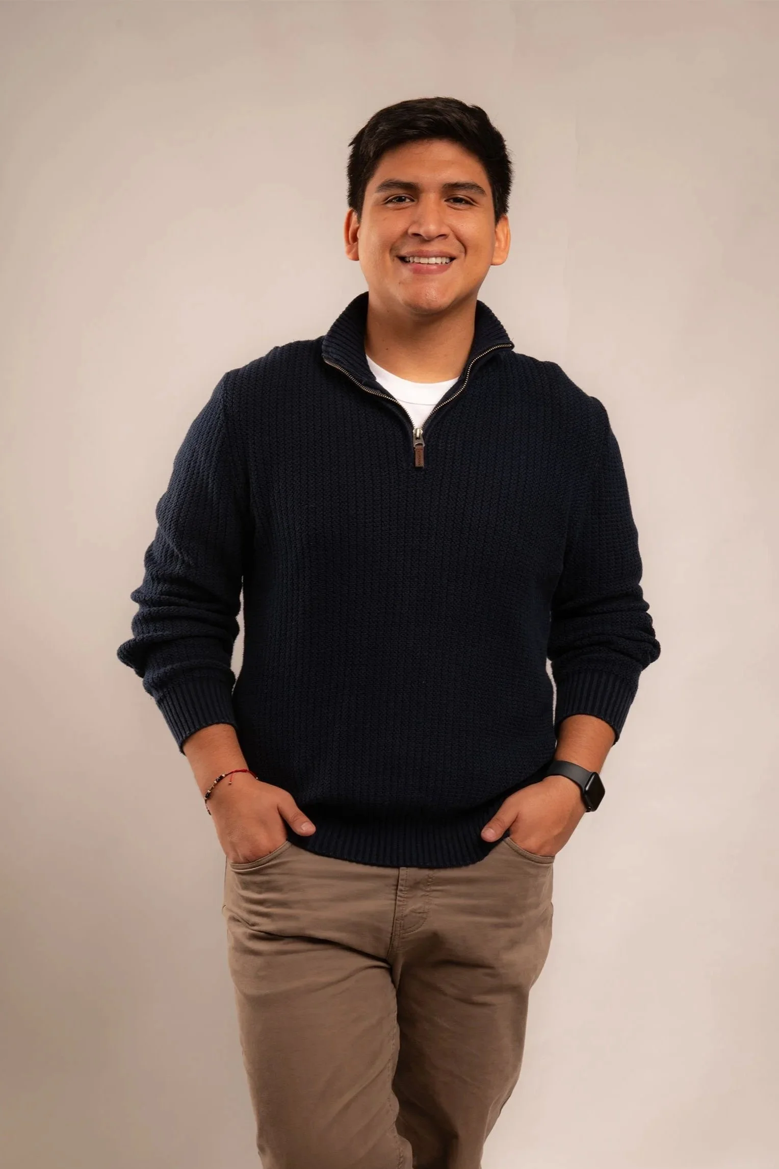 Joven hombre sonriendo, de cabello oscuro, con camiseta blanca y suéter negro con cremallera, pantalones beige, en fondo neutro.