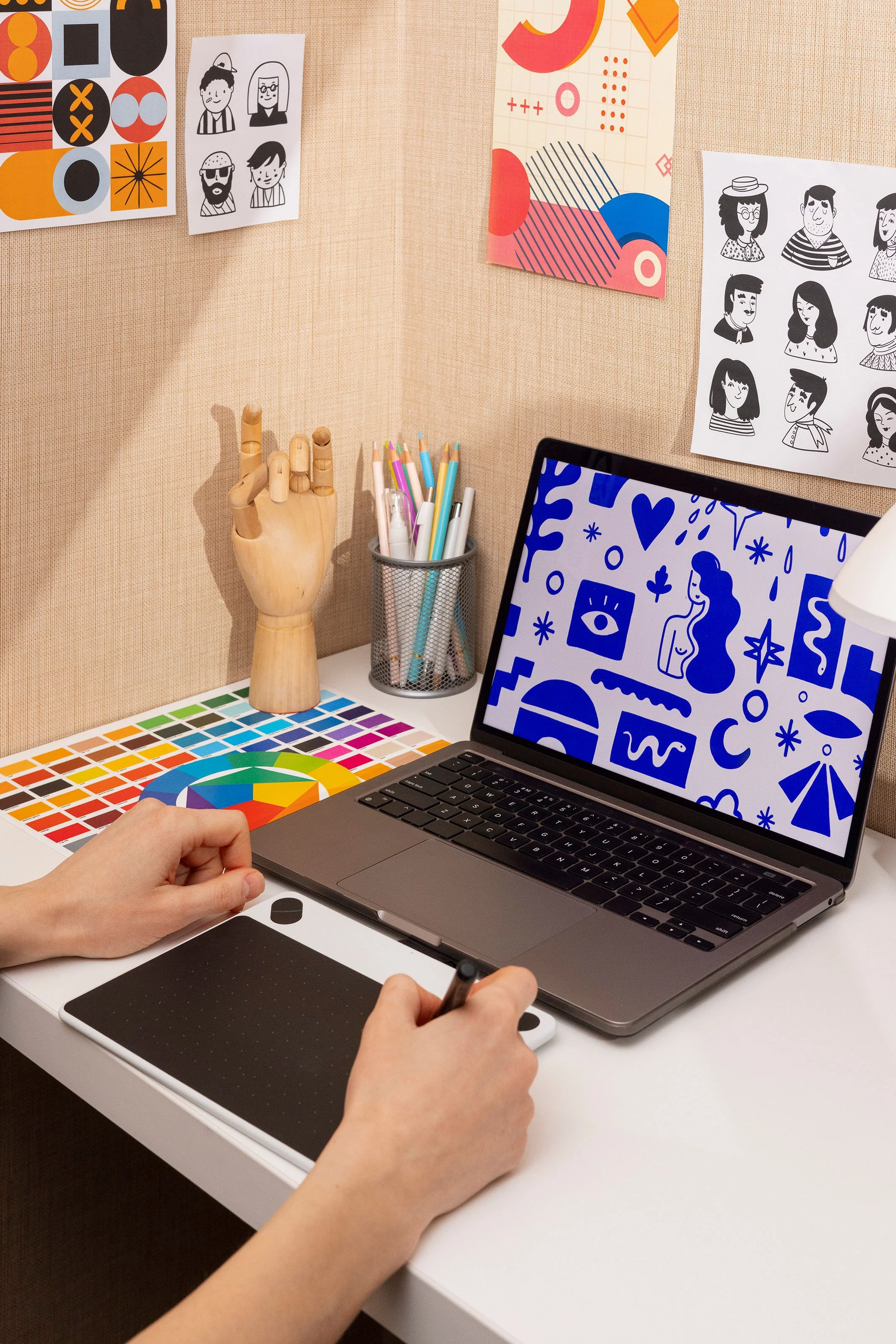 Espacio de trabajo con una mesa blanca, una tableta gráfica, una computadora portátil, un conjunto de lápices en un recipiente de malla, una figura de mano de madera para dibujo, y varias hojas y carteles con dibujos y diseños artísticos en la pared.