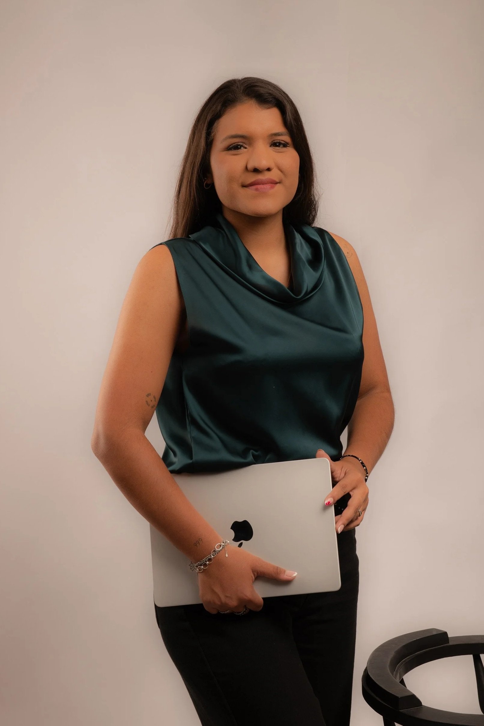 Mujer con cabello largo y oscuro, vistiendo blusa verde de seda y pantalones negros, sosteniendo una laptop plateada con manzana, de pie junto a una pared blanca, en un ambiente de oficina.