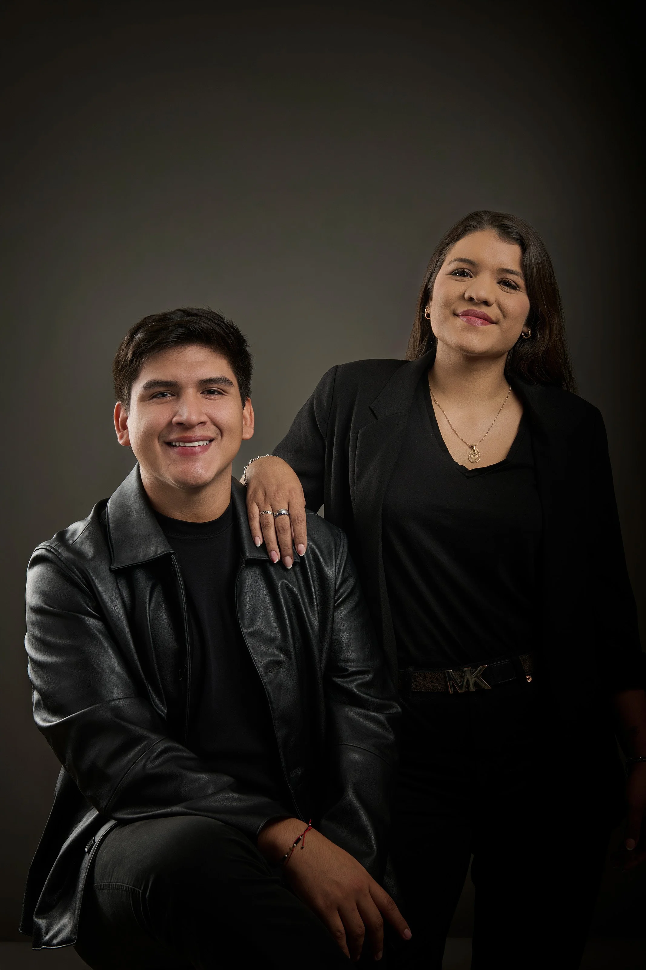 Un joven y una joven posando en un fondo oscuro. La joven lleva un blazer negro y un collar, el joven usa chaqueta de cuero negro y camiseta de color oscuro.