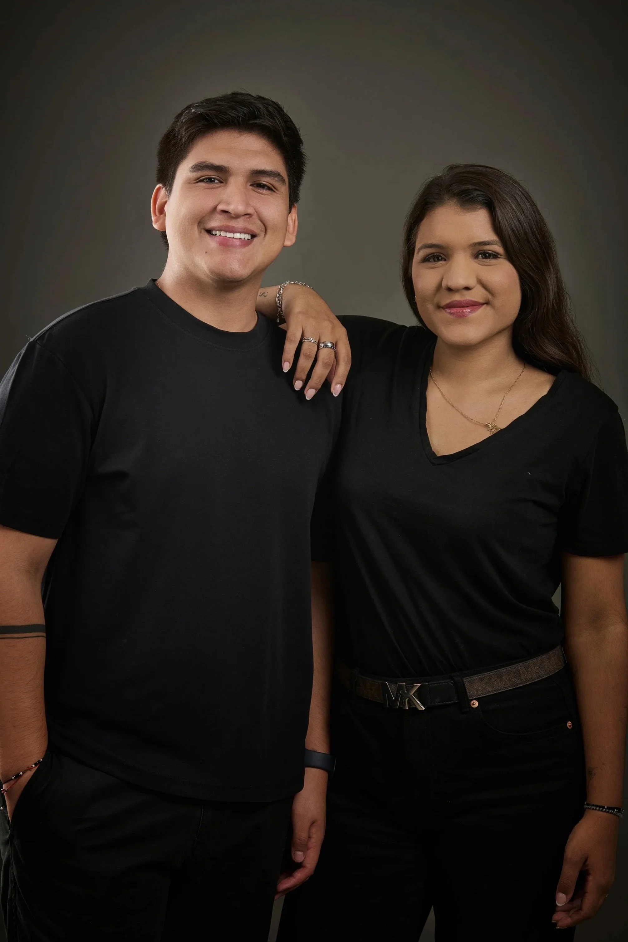 Dos personas, un hombre y una mujer, sonrientes, de brazos cruzados, frente a fondo gris oscuro, vestidas de negro.