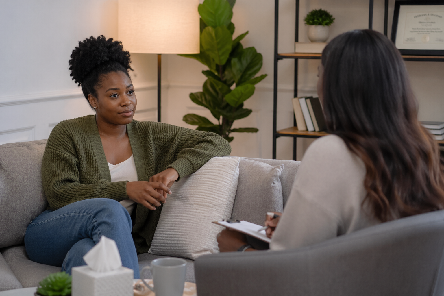 A woman with curly black hair sitting on a sofa having a conversation with a therapist in an office, with a lamp, plant, and framed certificate in the background.