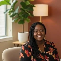 Smiling woman with braided hair sitting in a cozy room with a large potted plant, a window, and a table with a lamp.