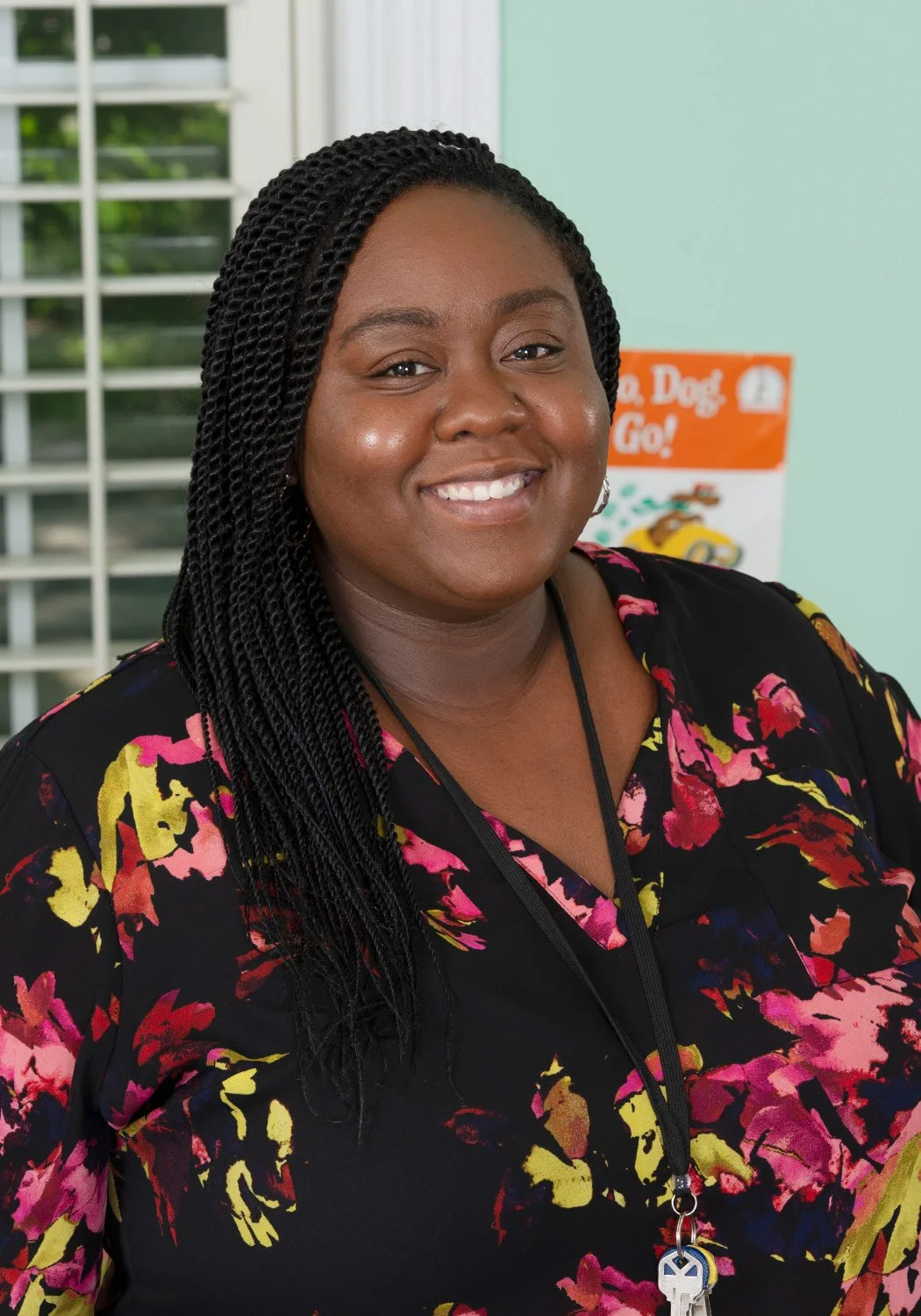 Smiling woman with braided hair wearing a floral dress, standing indoors near a window and a colorful poster.