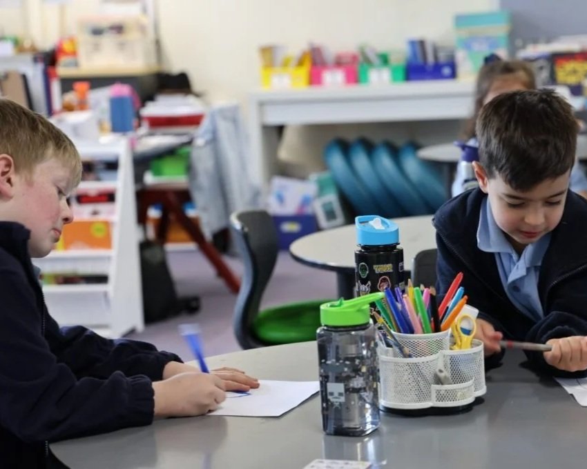 Two young boys sitting at a desk in a classroom, working on drawings. The desk has pencil holders with colorful markers, a water bottle, and scissors. Classroom supplies and shelves are visible in the background.