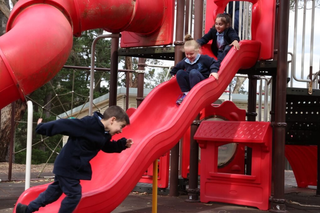 Three children playing on a red playground slide at a playground area with trees in the background.