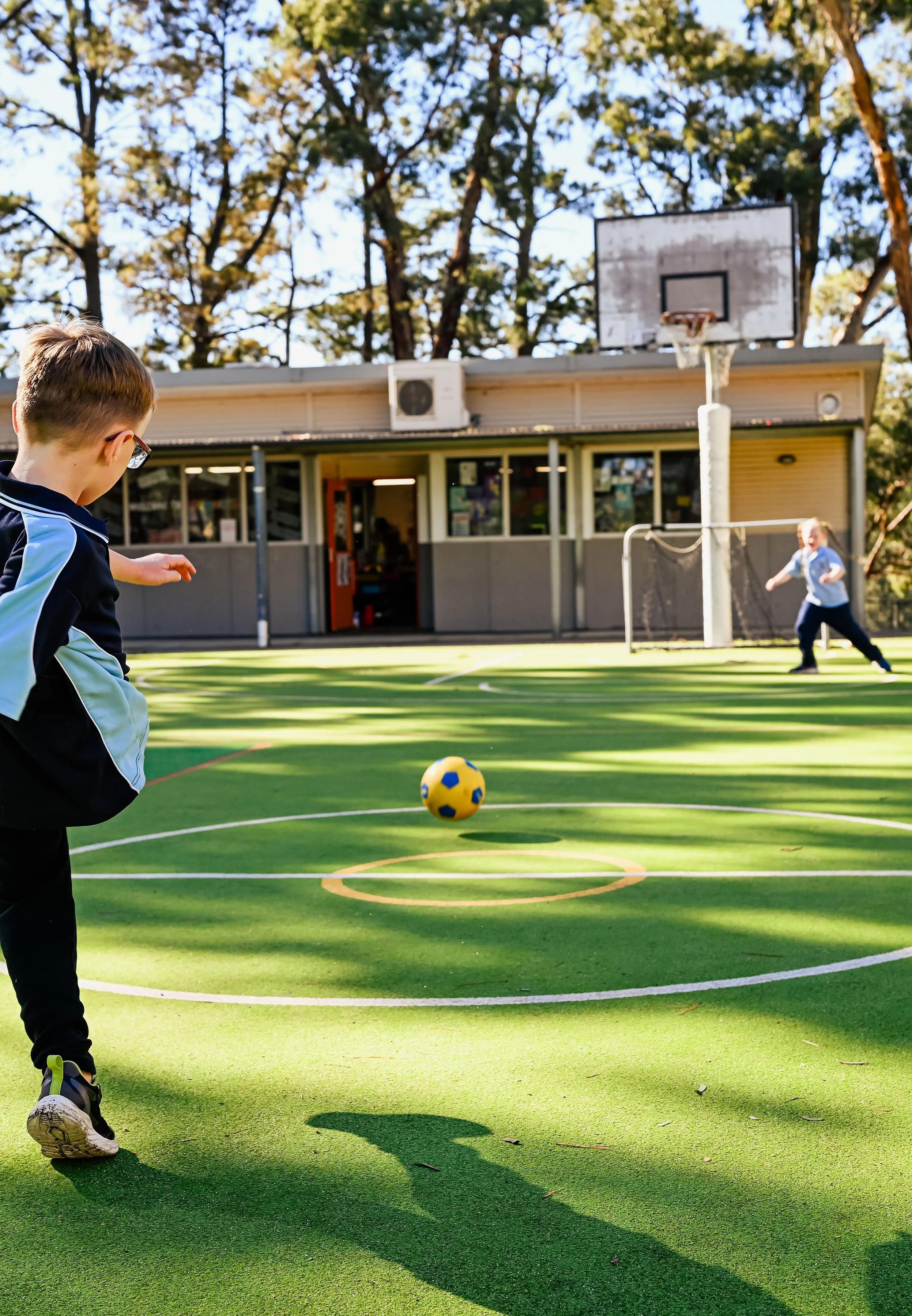 A young boy in a primary school sports uniform kicks a yellow and blue soccer ball on an outdoor green sports field under trees, with a basketball hoop and a building in the background.