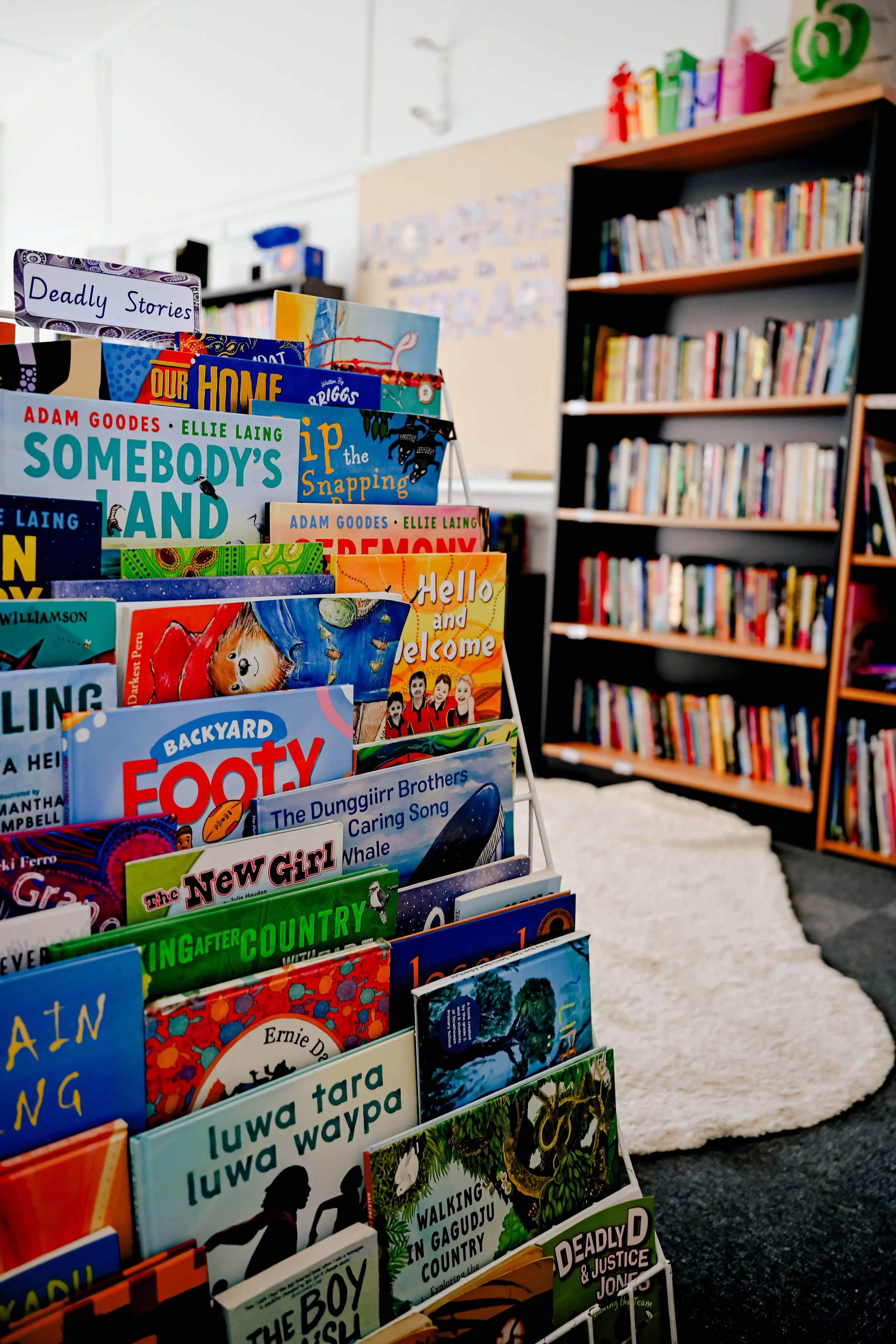 Close-up view of a bookshelf in a library filled with children's books with colourful covers and titles, against a blurred background of more bookshelves and a beige wall.
