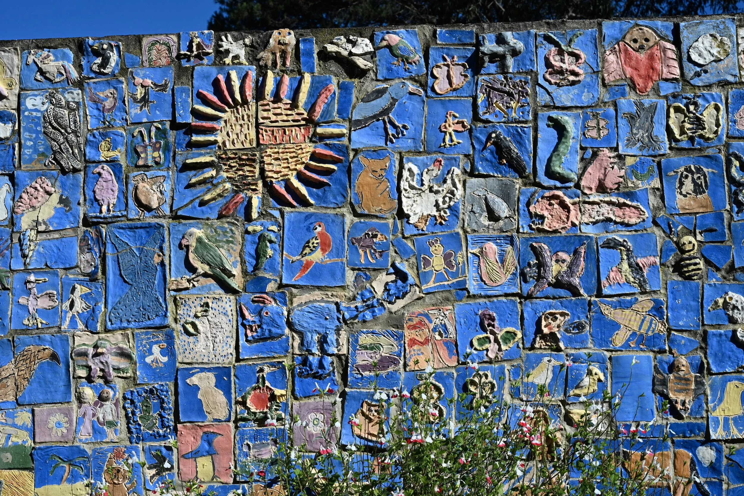 Colorful ceramic mosaic mural depicting various animals, insects, and patterns on a brick wall, with flowers and greenery at the bottom.