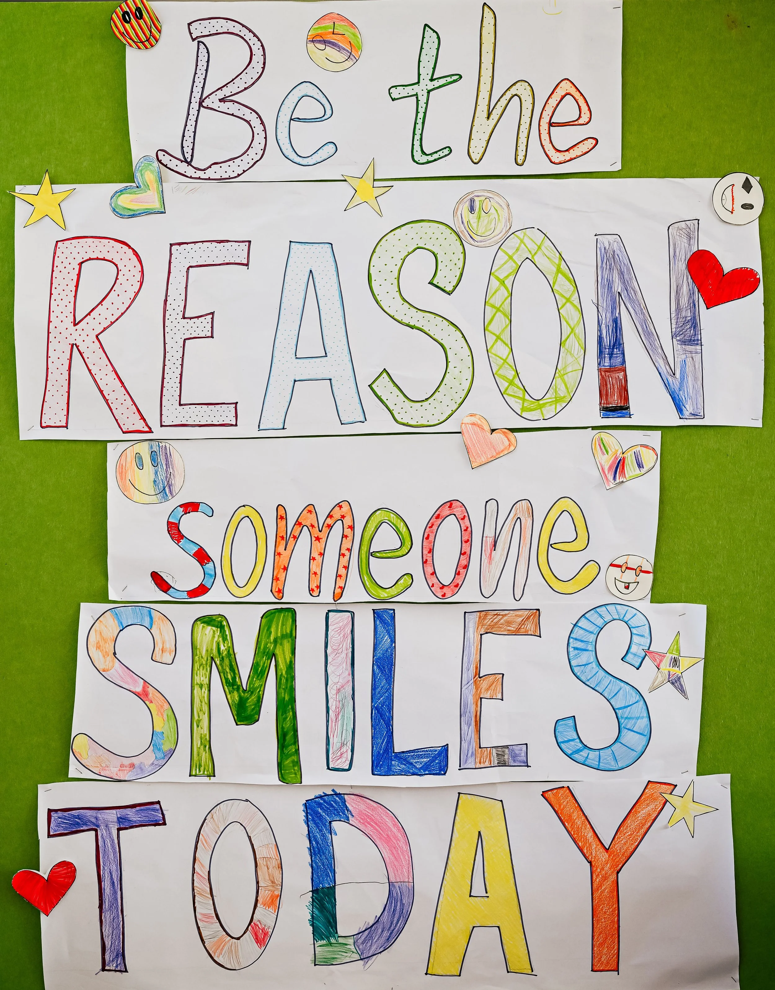 Colourful handmade posters displaying a messages that reads "Be the reason someone smiles today," with various doodles, stars, and hearts on a green background.
