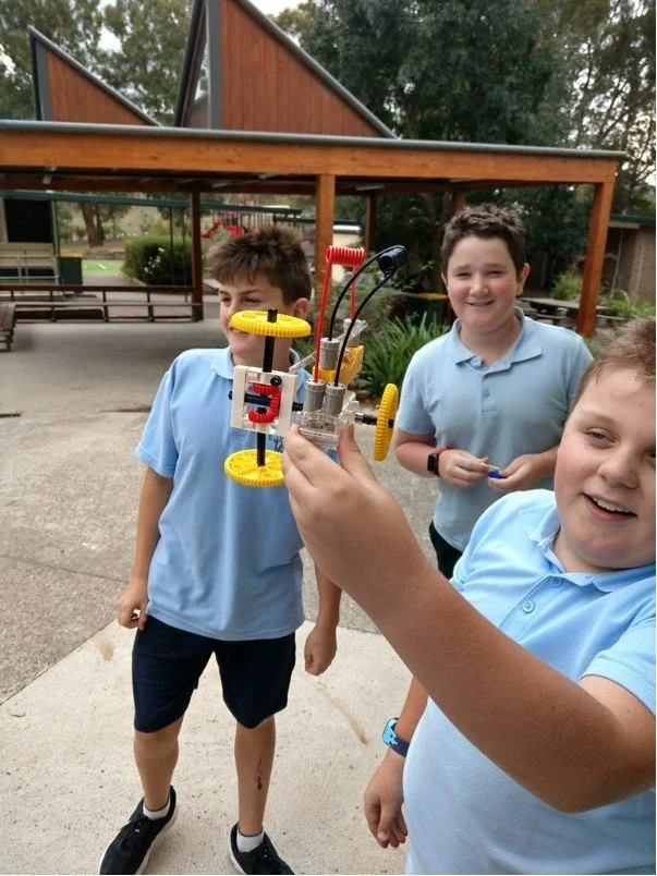 Three boys in blue school uniforms observing a small robotics project held by one of the boys outdoors at school.