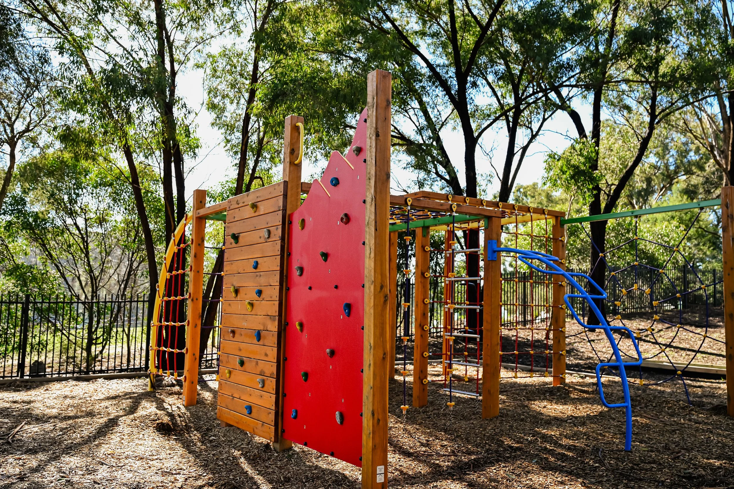 Colourful outdoor playground structure with climbing walls and ropes in a park with trees and a black fence.