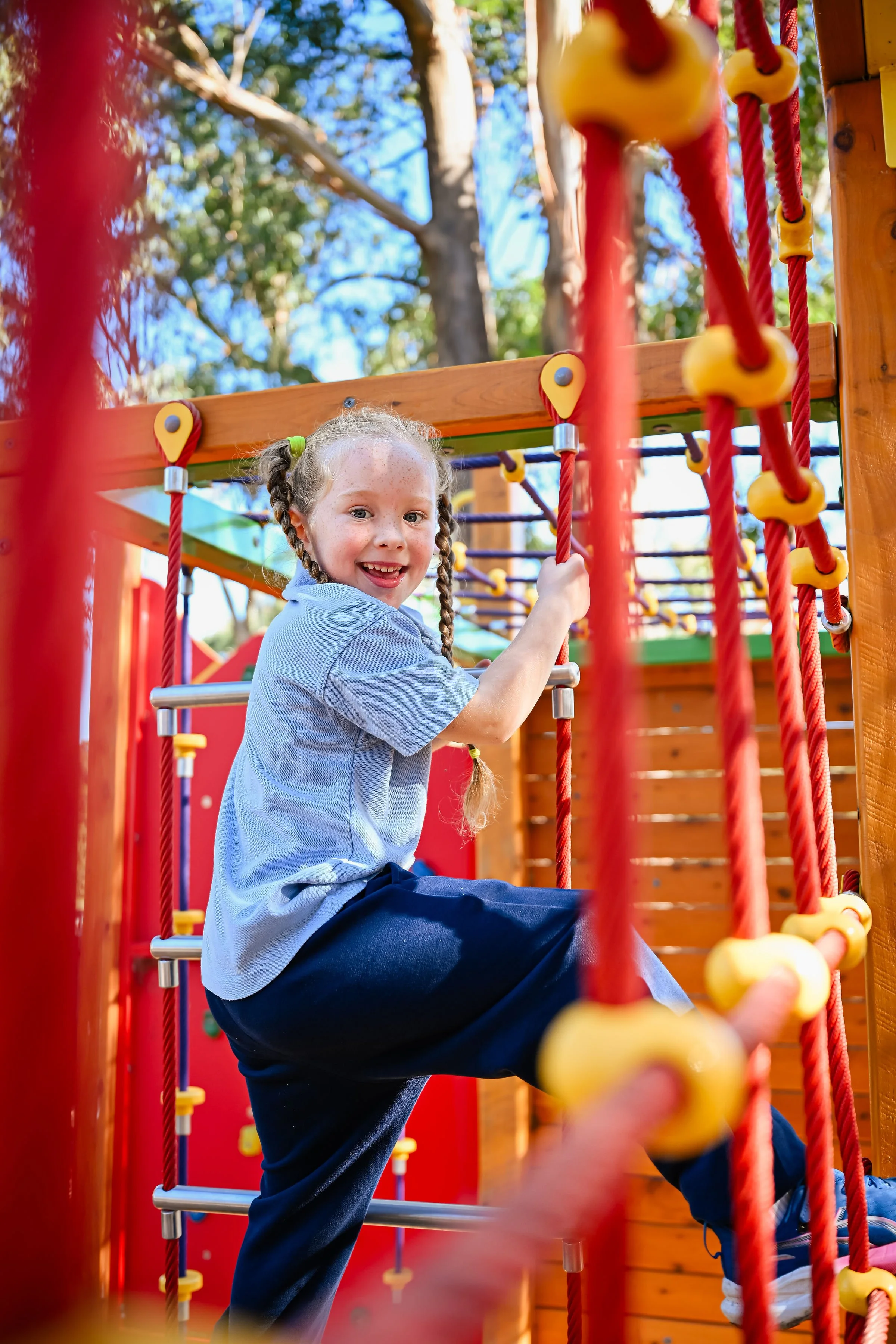 A young girl with braided hair and freckles enjoying herself on a climbing playground structure with red ropes and yellow connectors outdoors surrounded by trees.