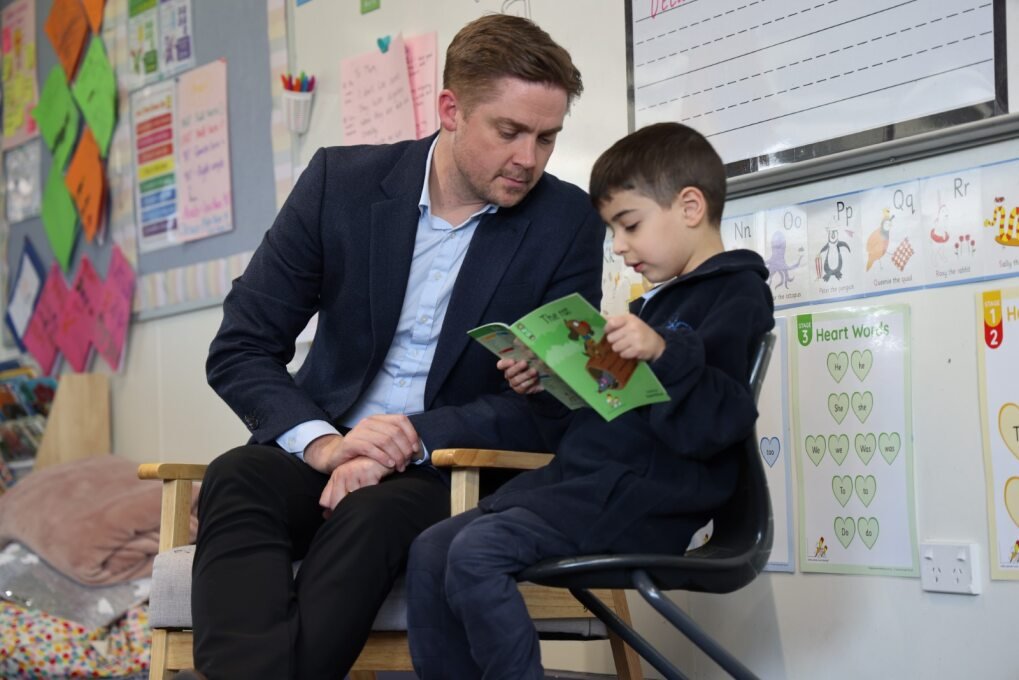 A man and a young boy sit closely together in a classroom, with the boy reading a green and brown book while the man looks on attentively.