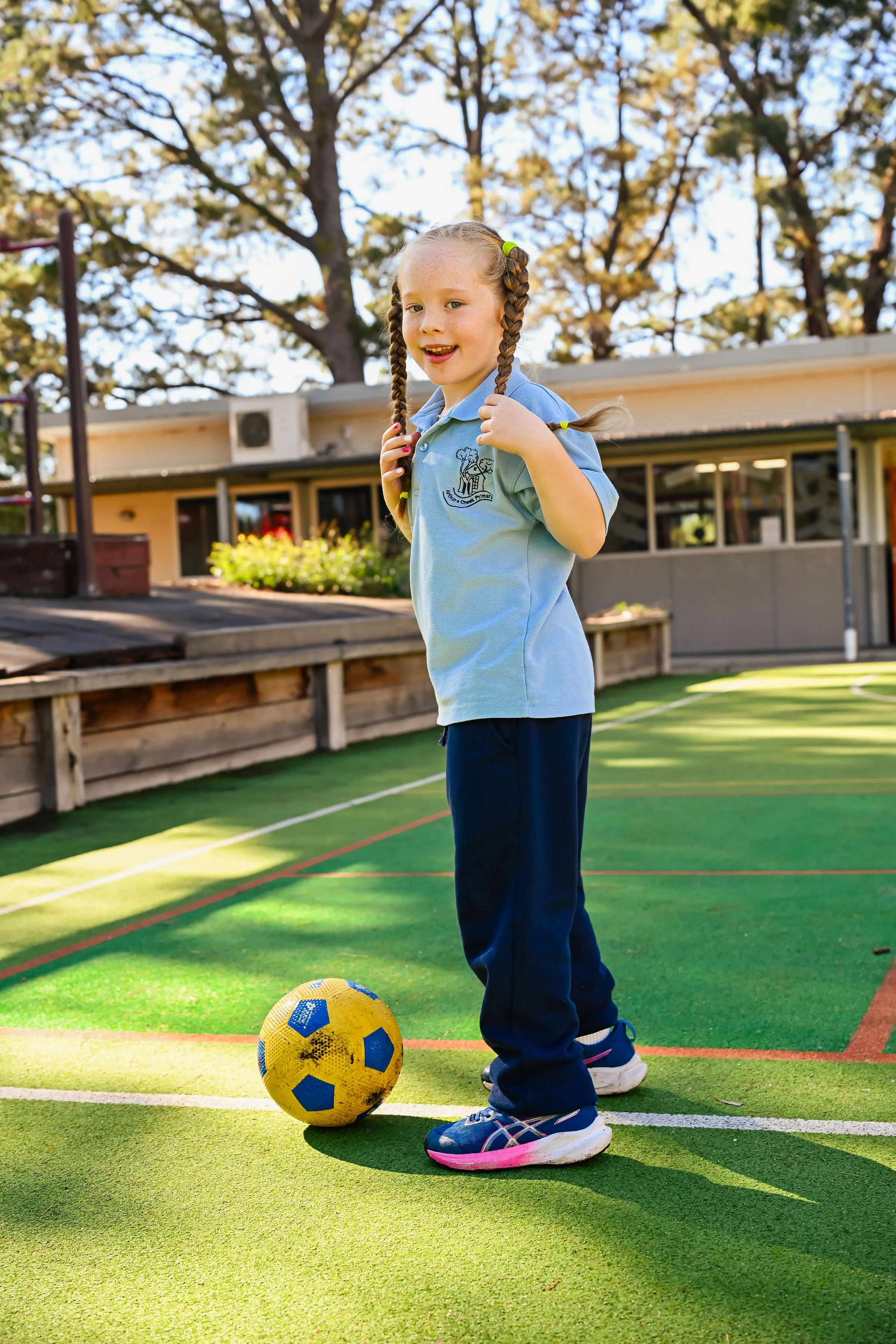 A young girl with braided hair stands on an outdoor sports field, holding a soccer ball with a yellow and blue design. She is wearing a light blue school uniform shirt and dark blue pants, with athletic shoes. There are trees and a building in the background, and the scene is sunny with clear sky.