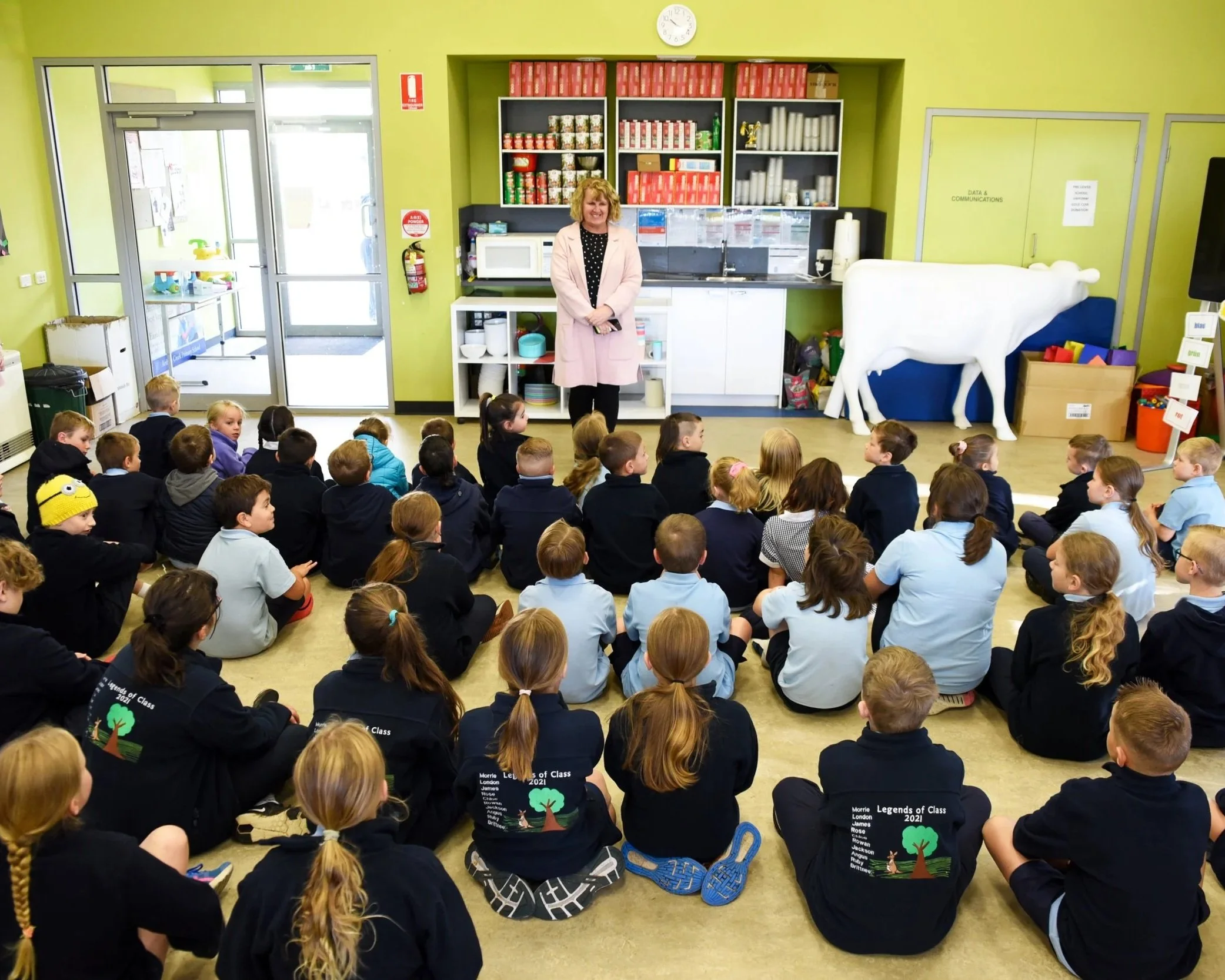 A teacher standing in front of a classroom full of students sitting cross-legged on the floor. The classroom has bright lime green walls, open shelving with books and supplies, and cartoon animal decorations. The students are wearing uniform shirts with a logo on the back, and some have colorful hair accessories.