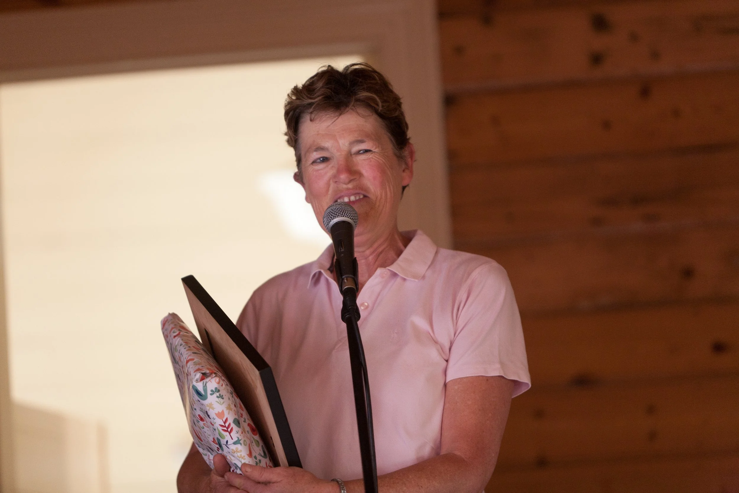 A woman with short curly hair, wearing a light pink shirt, is speaking into a microphone while holding a rolled-up piece of paper. She stands in front of a wooden wall and a bright background.