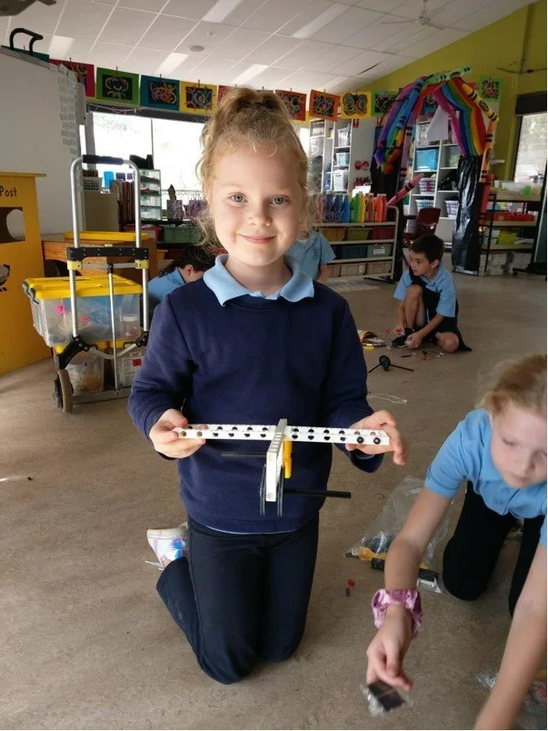 A young girl kneeling on the floor in a classroom, holding a small construction project made of plastic and metal, with other children in the background engaging in activities.