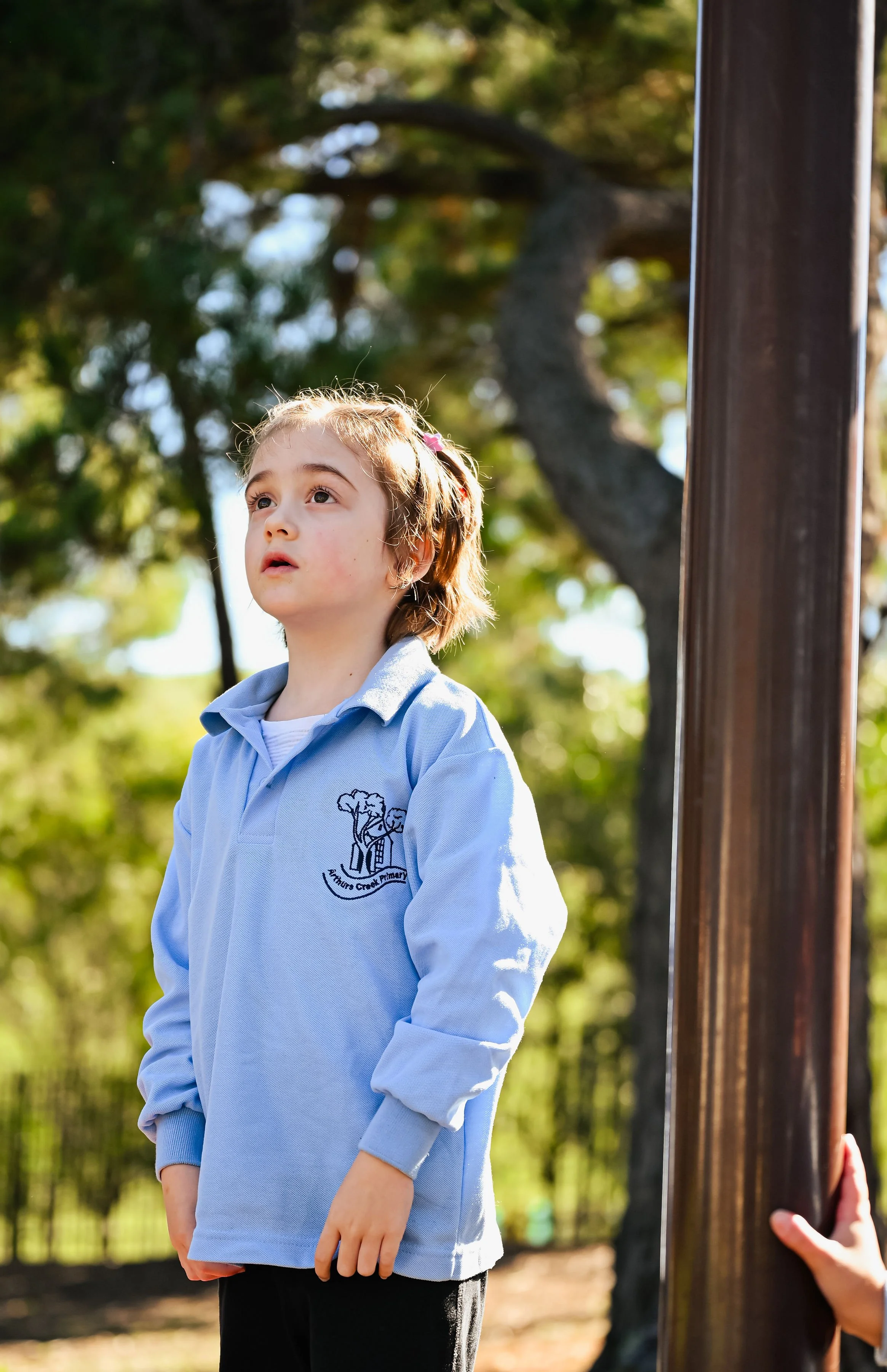 A young girl with light brown hair and a pink hair clip standing outdoors beside a dark brown pole, looking up with a curious expression. She is wearing a light blue school uniform with a logo on the chest and black pants. The background features green trees and sunlight filtering through the leaves.