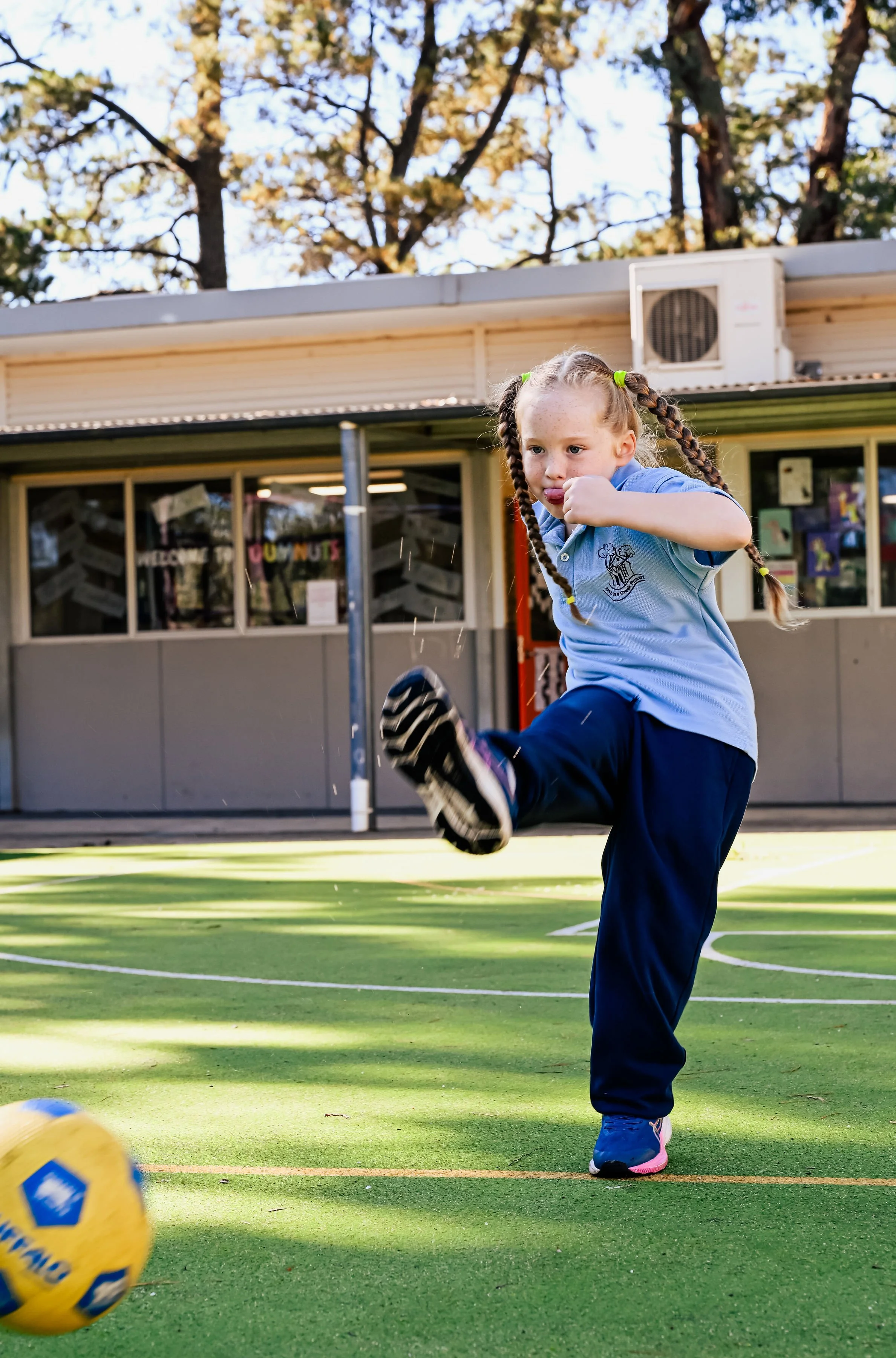 A young girl in a blue school uniform is kicking a yellow soccer ball on a green field during daytime. She has braided hair and is focused on the ball, with a building and trees in the background.