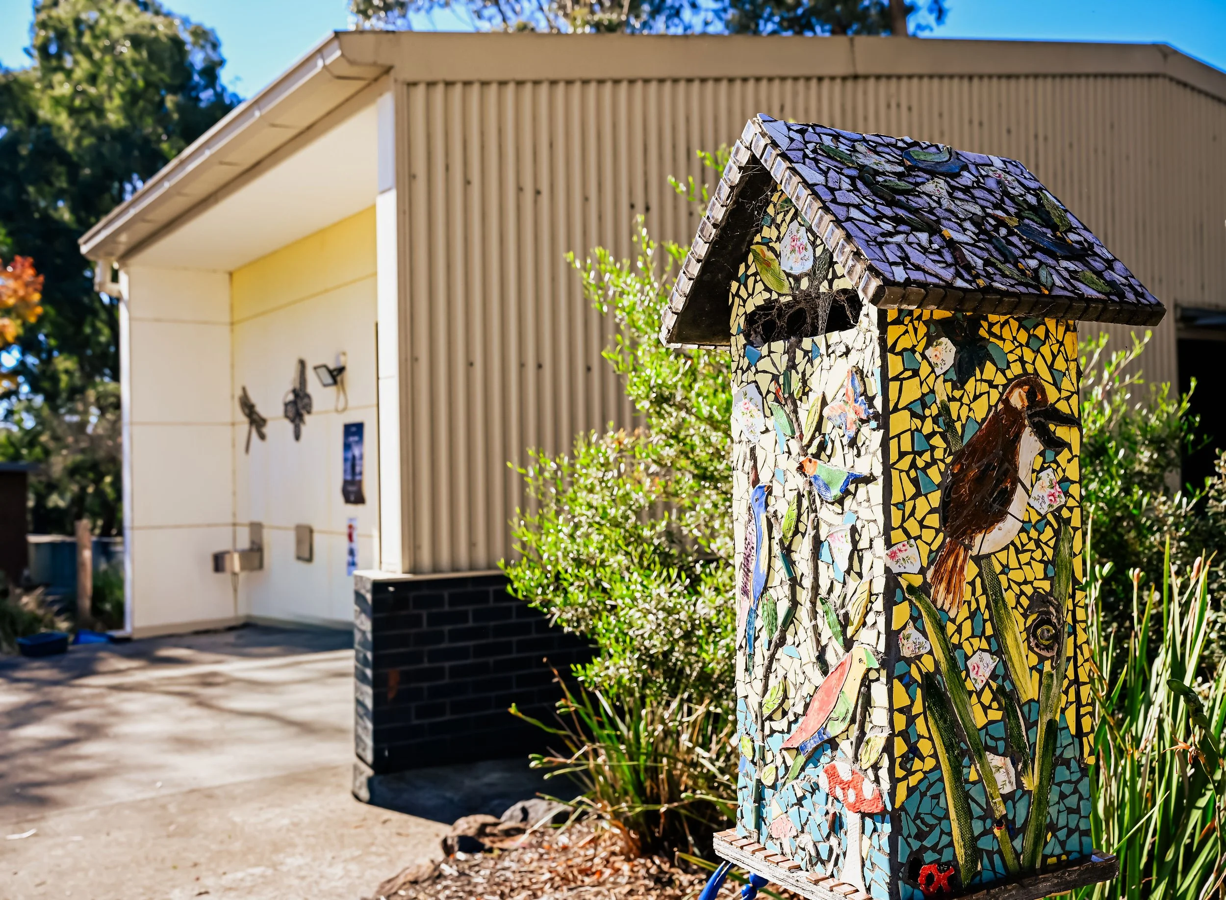 Colorful mosaic birdhouse in front of a building with beige siding and a white roof, surrounded by greenery.