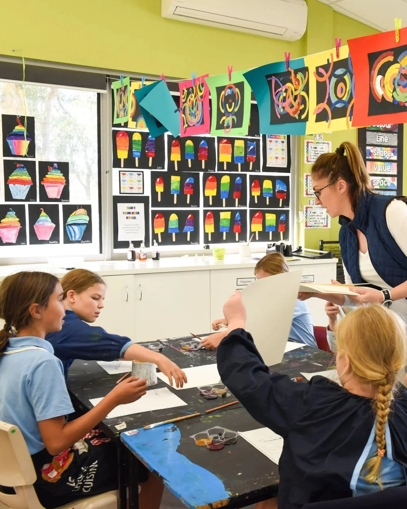 Kids in an art classroom working on paintings, with colorful artwork displayed on the walls and hanging decorations.