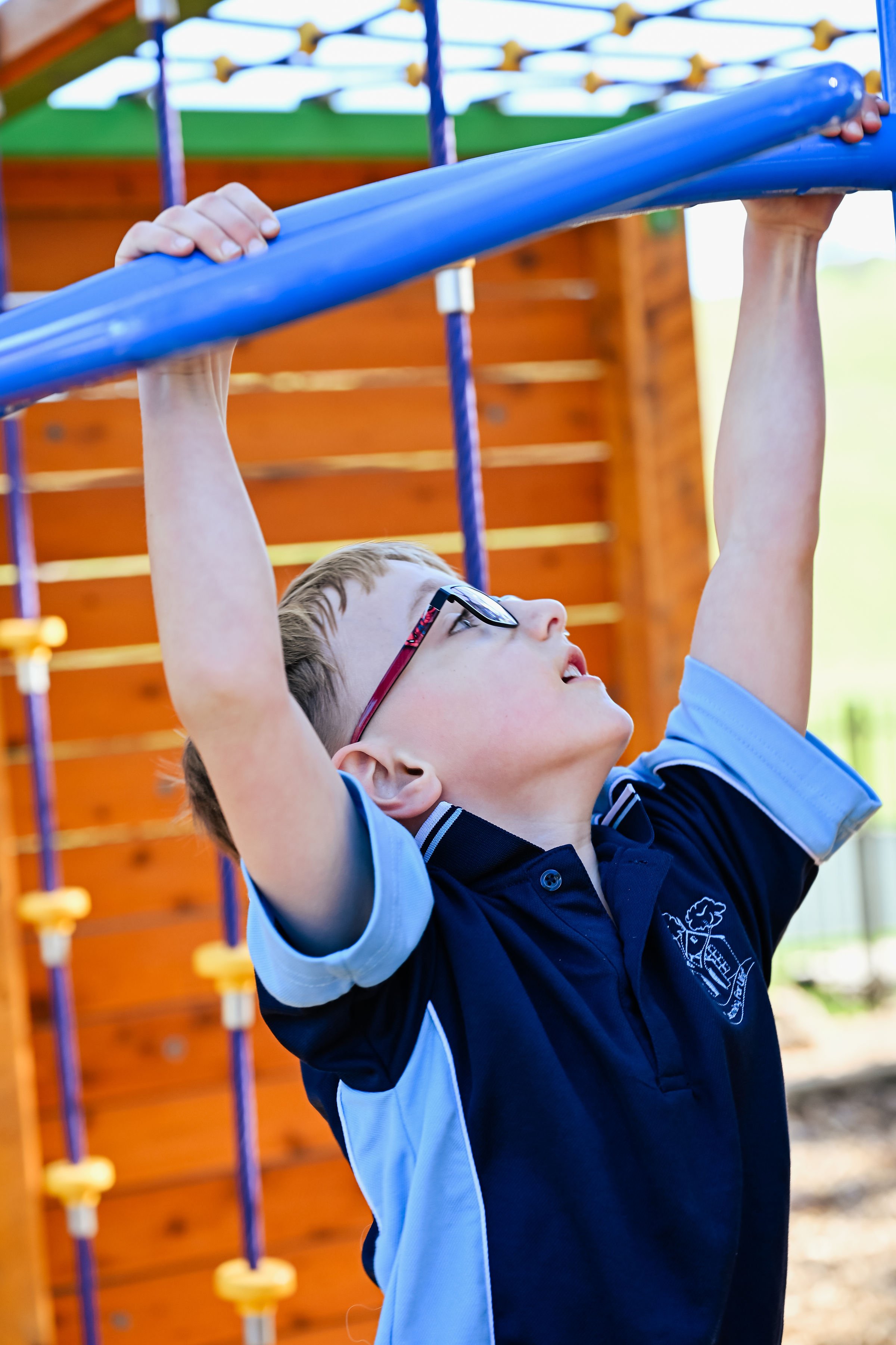 A young boy wearing glasses and a blue and black school uniform is hanging from a blue horizontal bar at a playground, with a wooden climbing structure in the background.