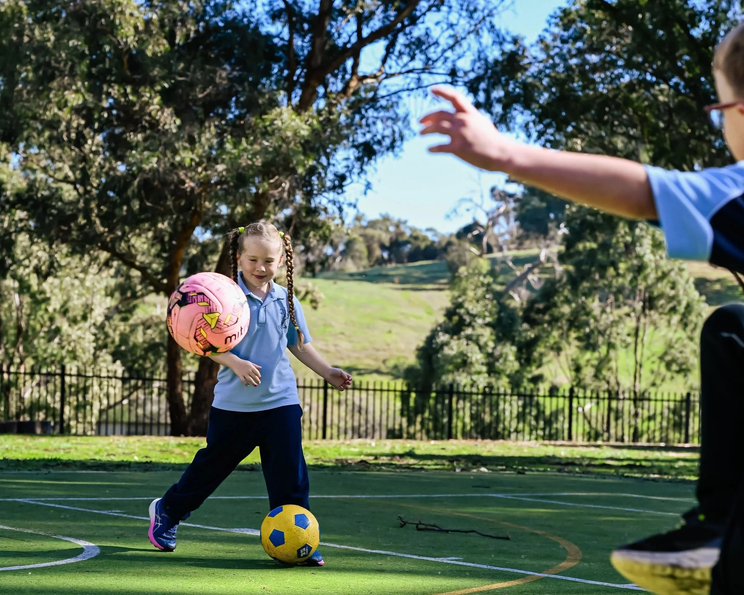 A young girl with braided hair wearing a blue shirt and navy pants, playing soccer on a green outdoor court, kicking a yellow and blue soccer ball with her right foot. Another child is reaching an arm out into the frame.,