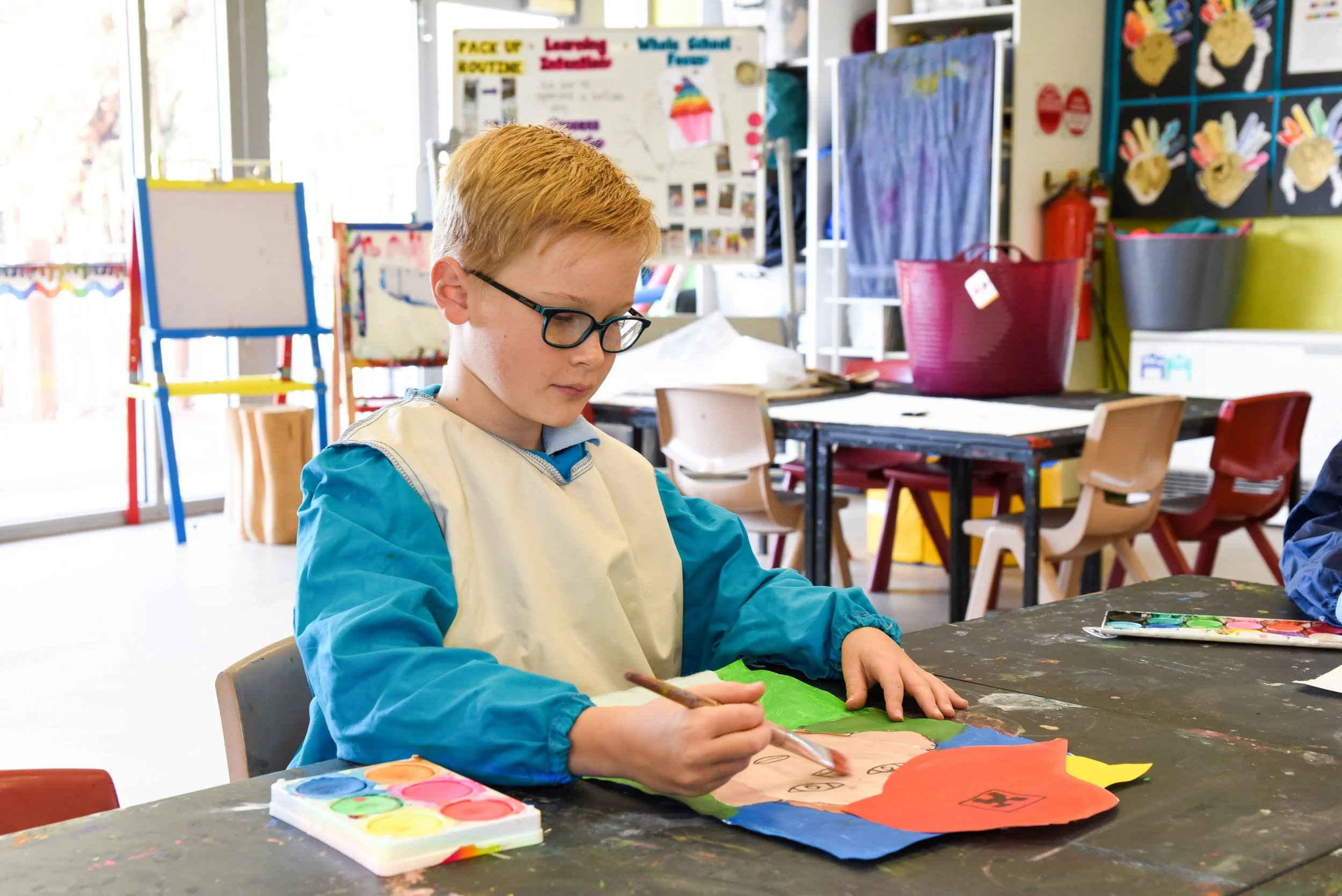 A young boy with red hair and glasses sitting at a table in an art classroom is painting. There are colourful supplies and artwork in the background.