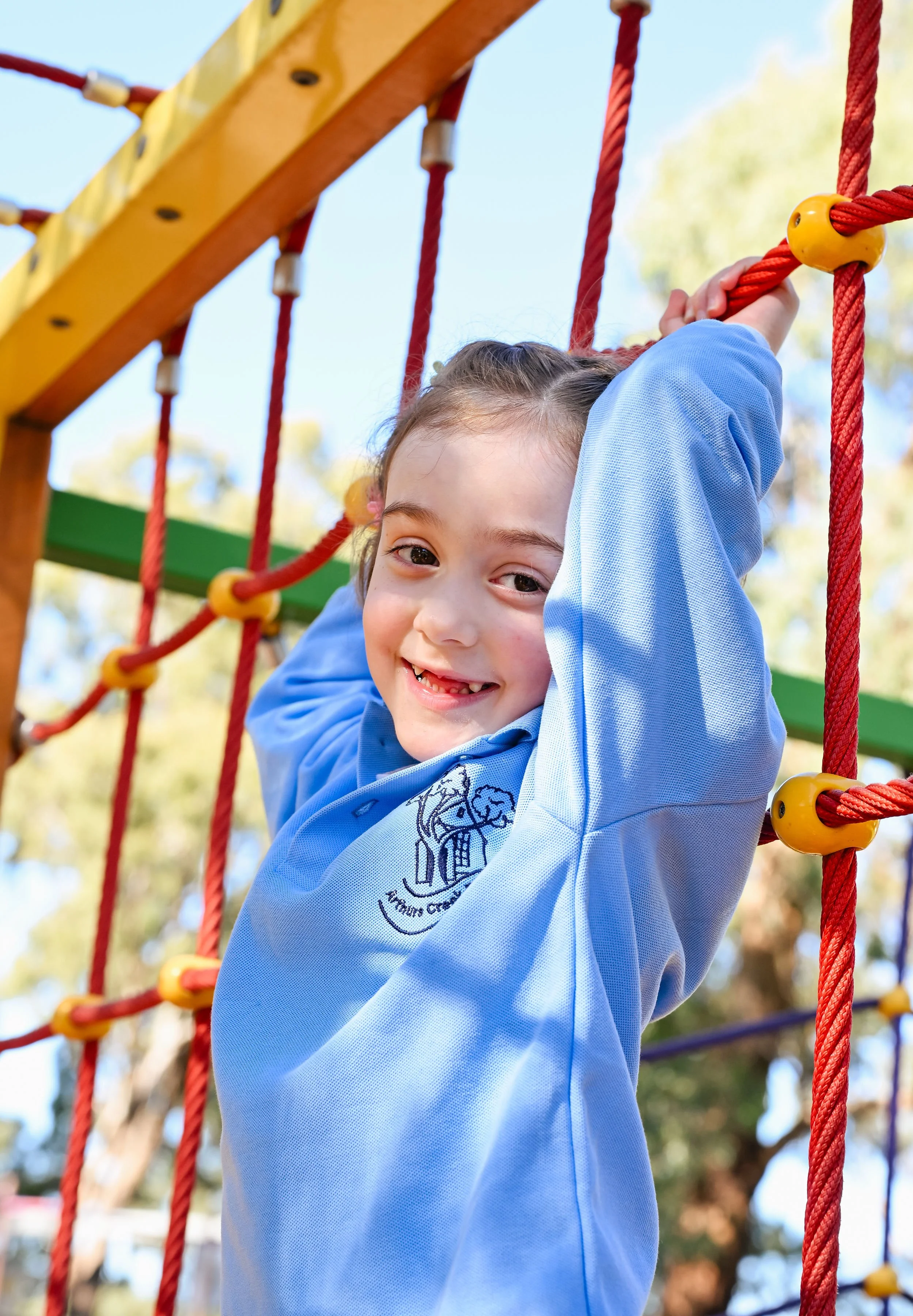 A young girl with brown hair and a blue long-sleeve shirt smiling and hanging on a red climbing rope at a playground, with trees and a blue sky in the background.