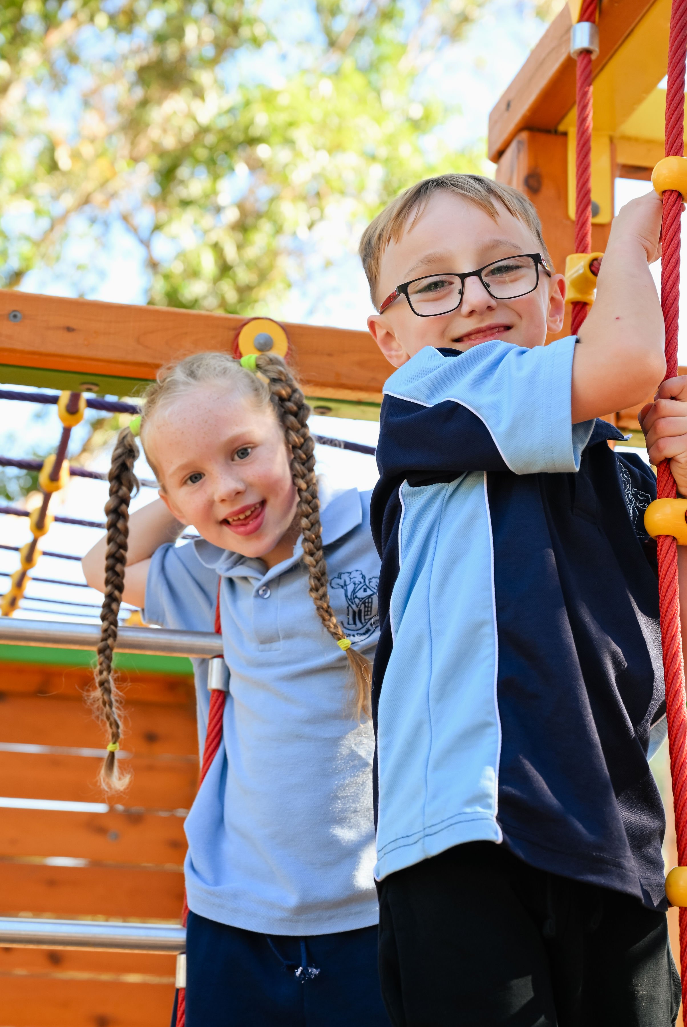 Two children, a girl and a boy, are on a wooden playground structure, climbing a red rope. The girl has braided hair and is smiling at the camera, while the boy is wearing glasses and also smiling. There are trees with green leaves in the background and sunlight shining through.