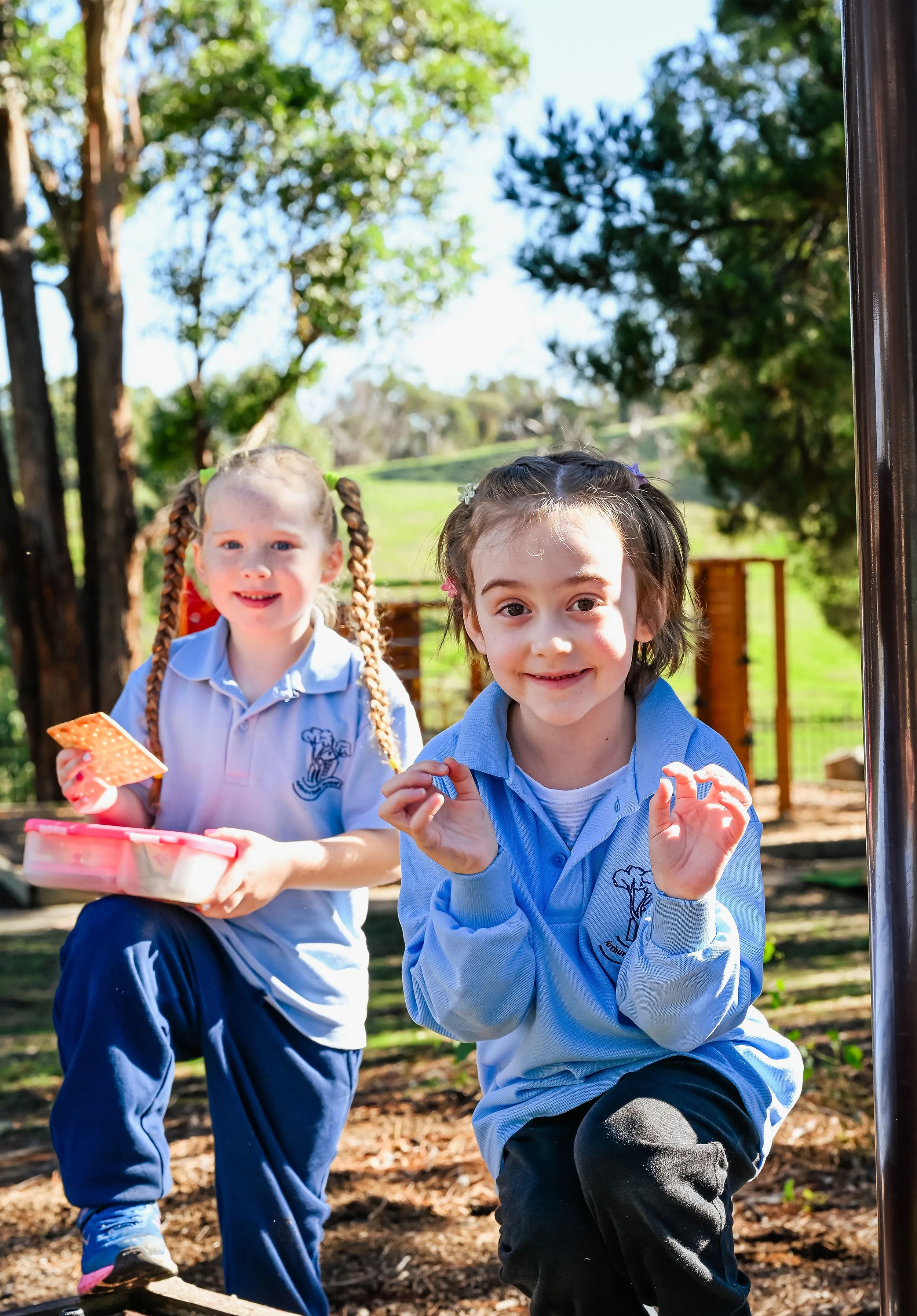 Two young girls in school uniforms at a park, smiling and standing near a playground structure.