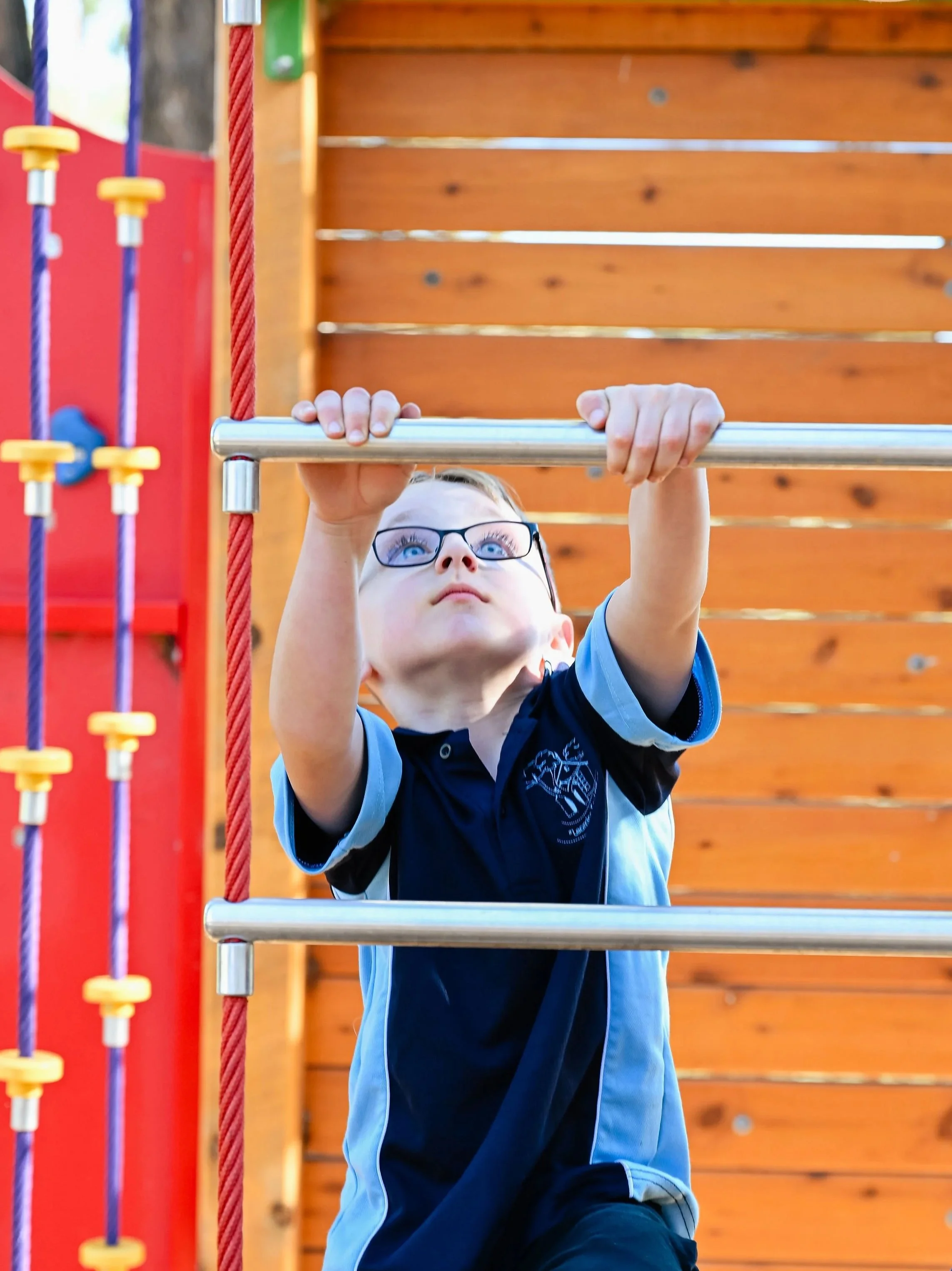 Child wearing glasses and a blue and black sports uniform hanging from a metal horizontal bar at a playground.