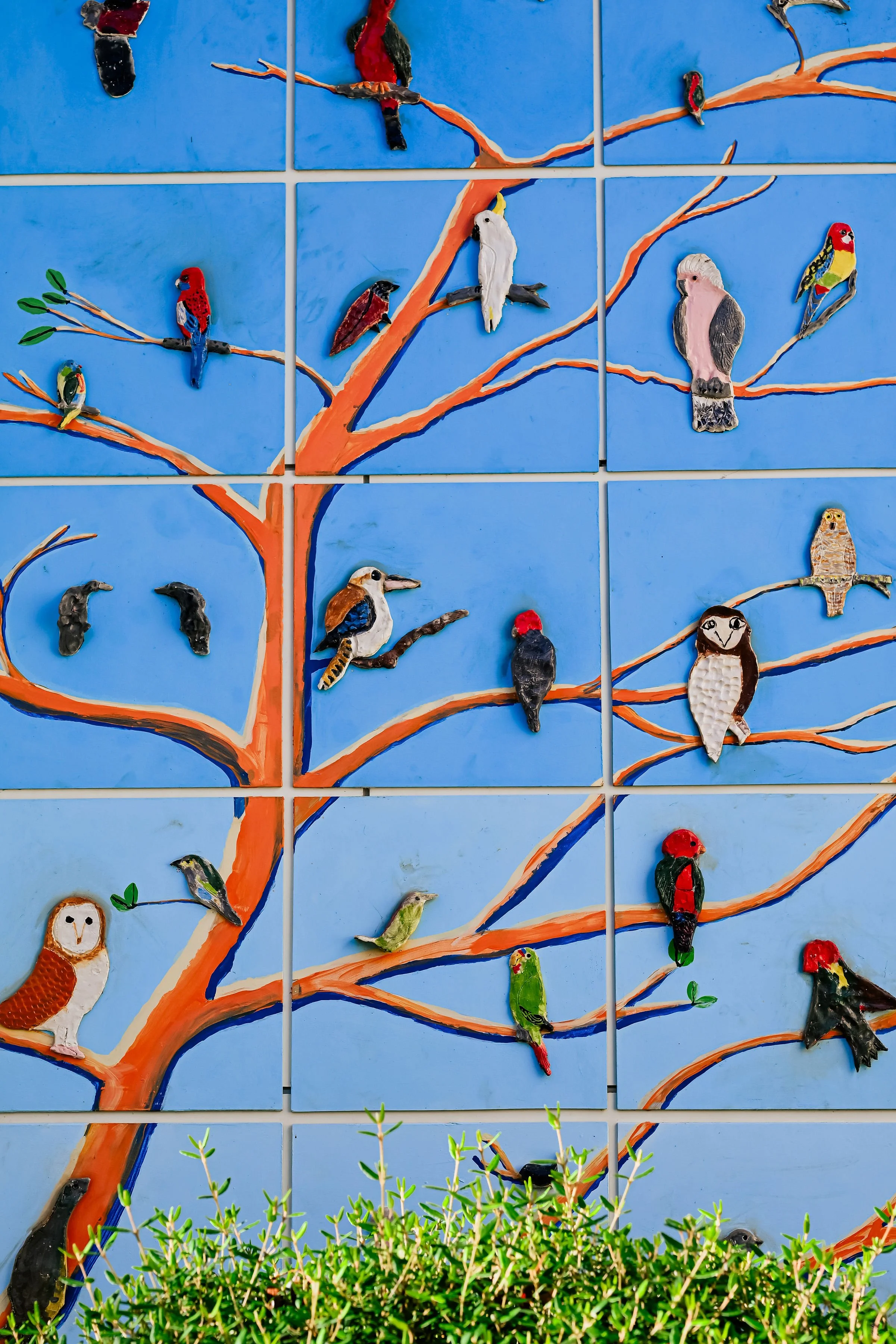 Colorful ceramic bird art on a blue tree mural with green shrub in front