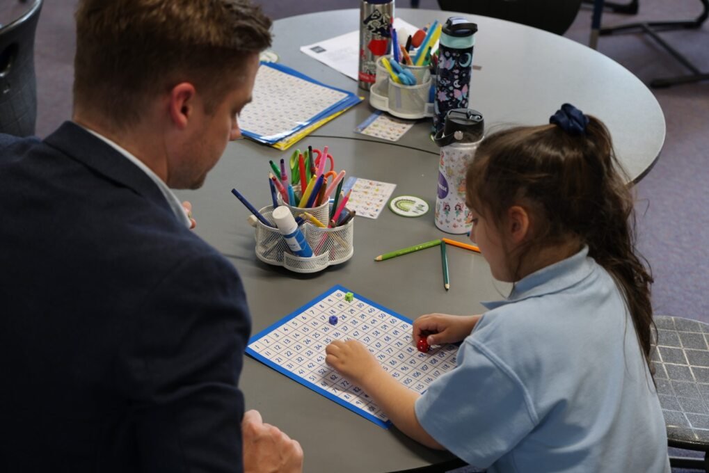 A man and a young girl playing a board game with dice at a table in a classroom or activity room.