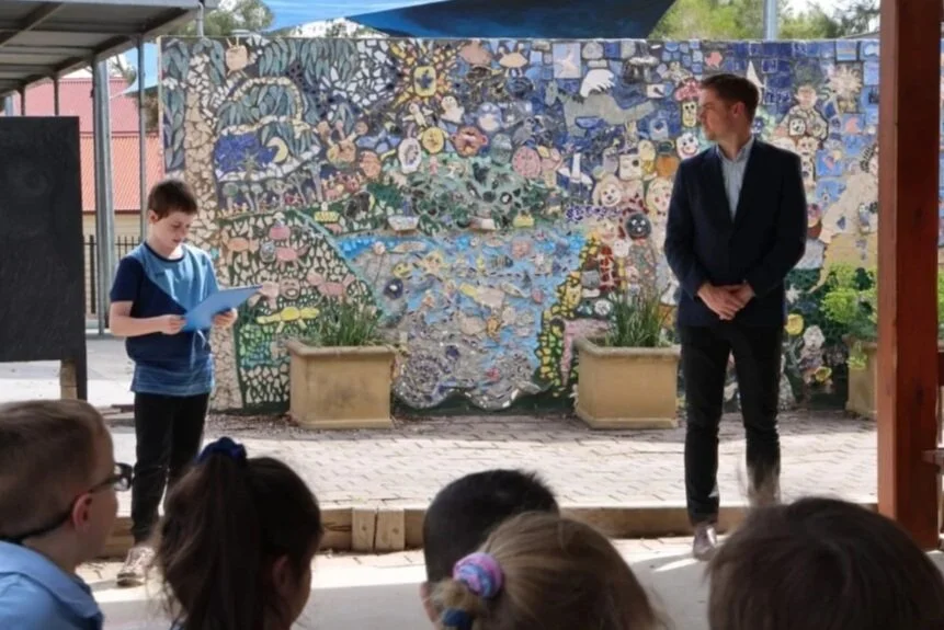 A young boy reads from a paper in front of an audience of children, with a man standing nearby, at an outdoor event with a colorful mosaic wall in the background.