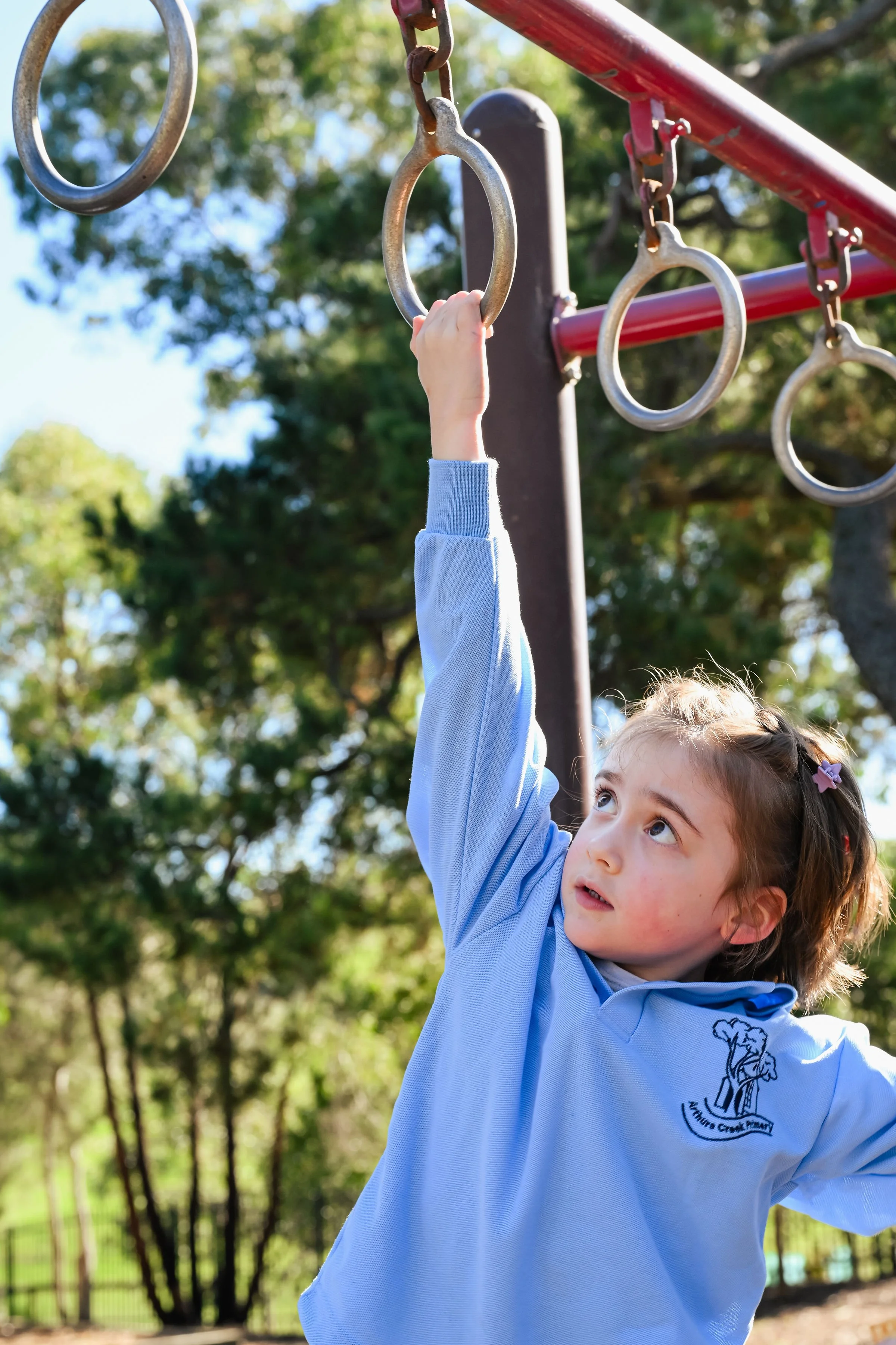 A young girl reaching up to grasp a metal ring on a playground structure outdoors, with trees and sunlight in the background.