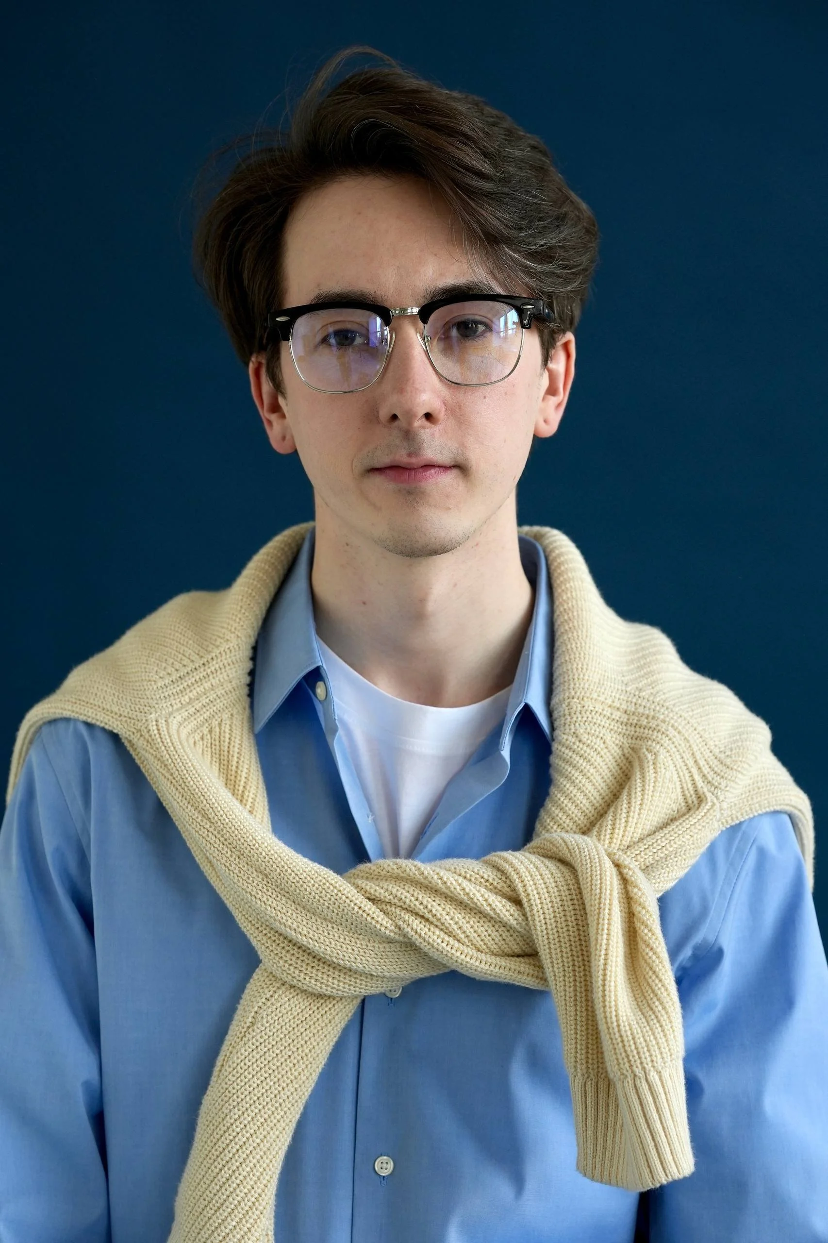 A young man with dark hair, wearing glasses, a light blue shirt, and a beige sweater draped over his shoulders, standing against a dark blue background.