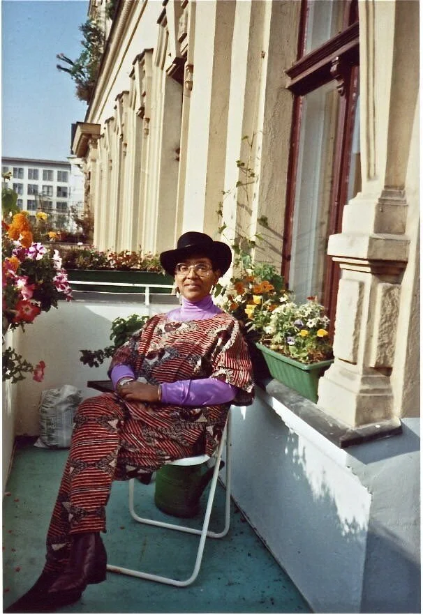 Audre Lorde sitting on a balcony adorned with colorful flowers, wearing a patterned dress, purple shirt, and a black hat, with a city building in the background.