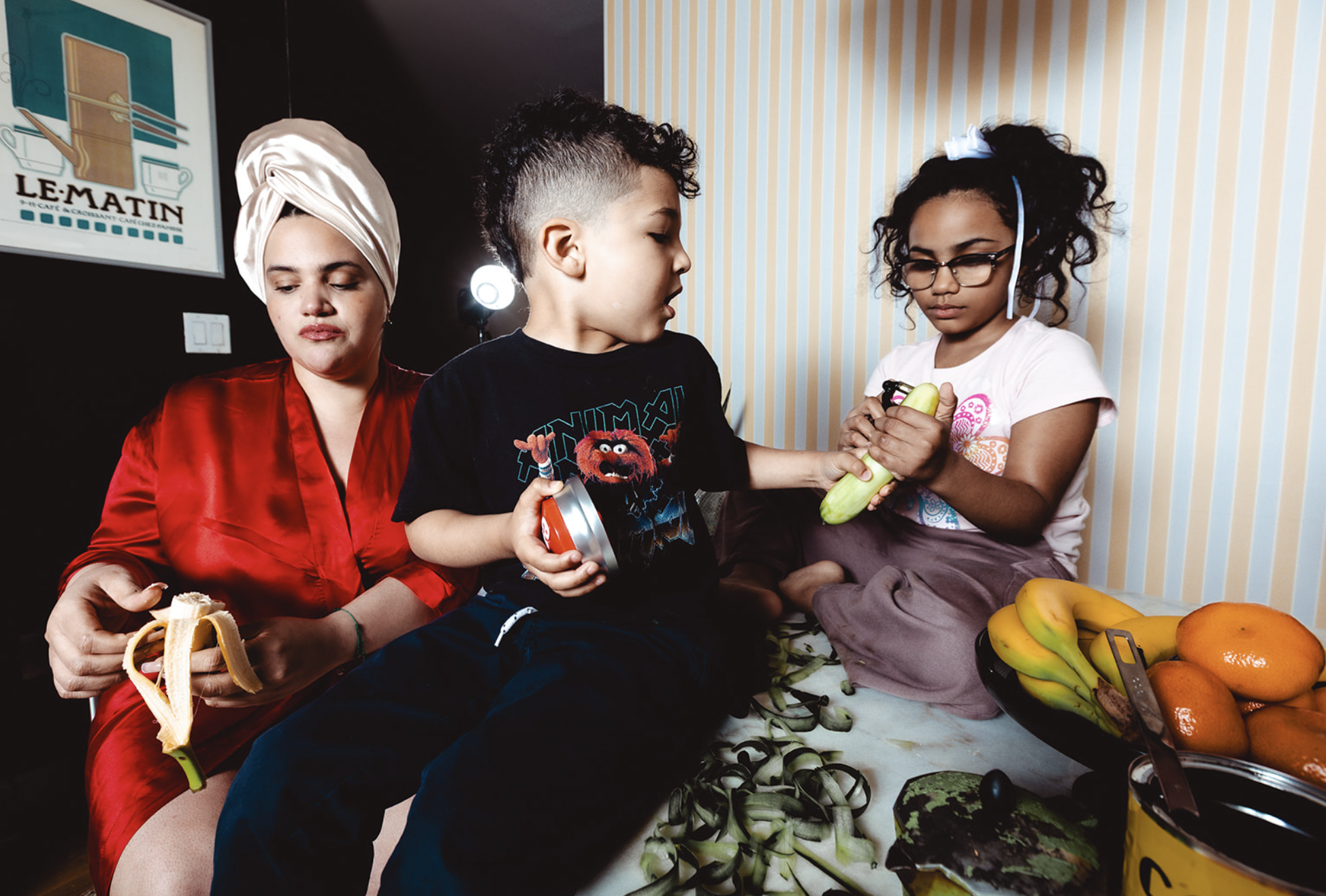 A woman in a white headwrap and red dress sitting with two children, a boy and a girl, who are examining a cucumber in a room with striped wallpaper. A table with bananas, oranges, and other fruits is in front of them.